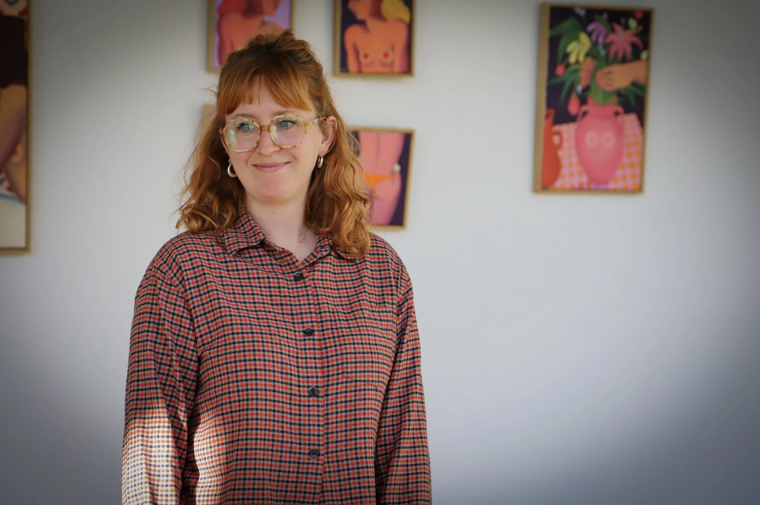 A woman with red hair, glasses, and hoop earrings is smiling and standing in an art gallery with colorful paintings of abstract and floral designs behind her.