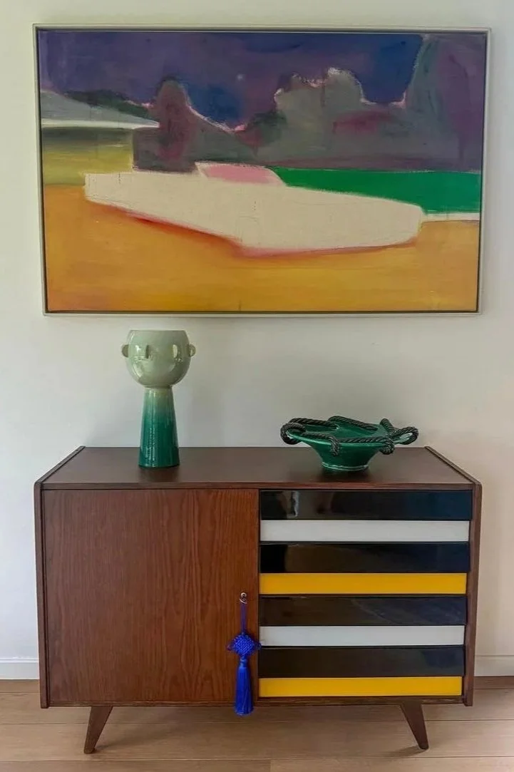 A wooden sideboard with a tall green vase and a green bowl on top, hanging on white wall, with a colorful abstract painting above.