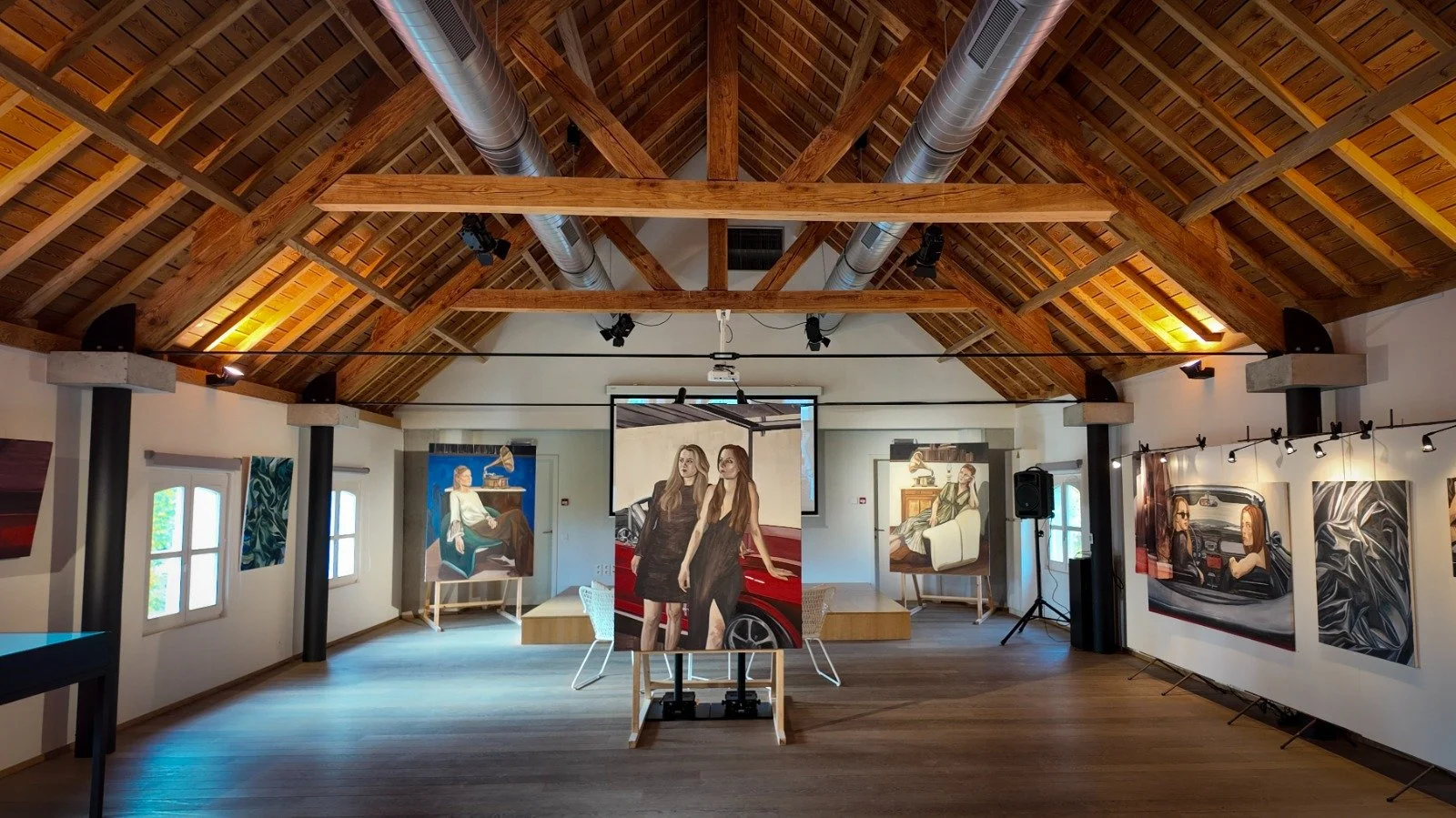 Interior of an art gallery with wooden vaulted ceiling and artwork on white walls, including paintings of women and abstract designs.