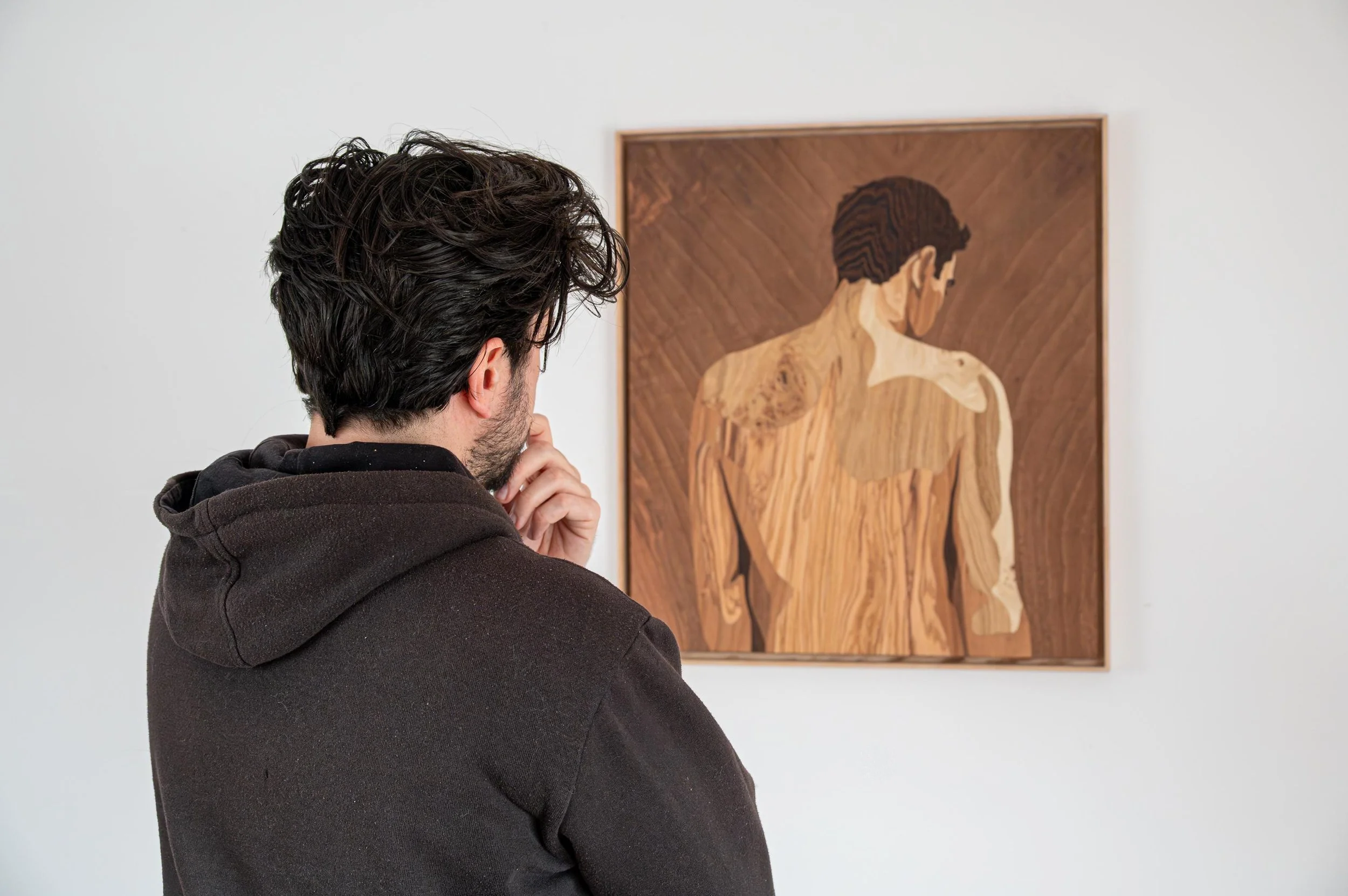 A man with dark, curly hair and a beard, wearing a dark hoodie, looking at a wooden abstract art piece of a man's back with short, dark hair, on a white gallery wall.