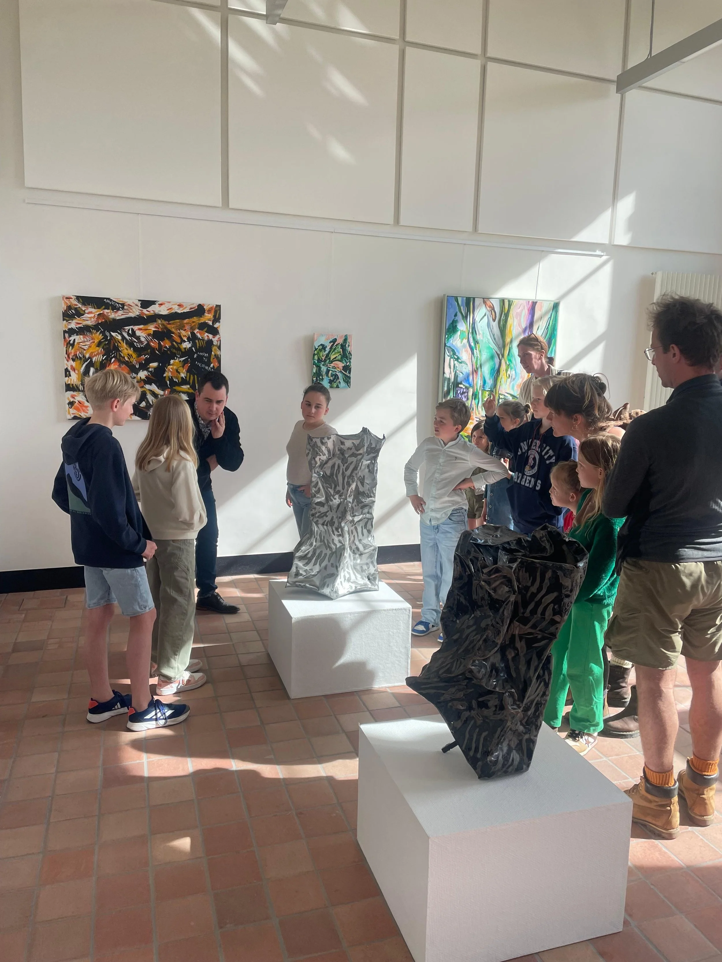 People, including children and adults, observing artwork in an art gallery with paintings on the wall and sculptures on white pedestals.