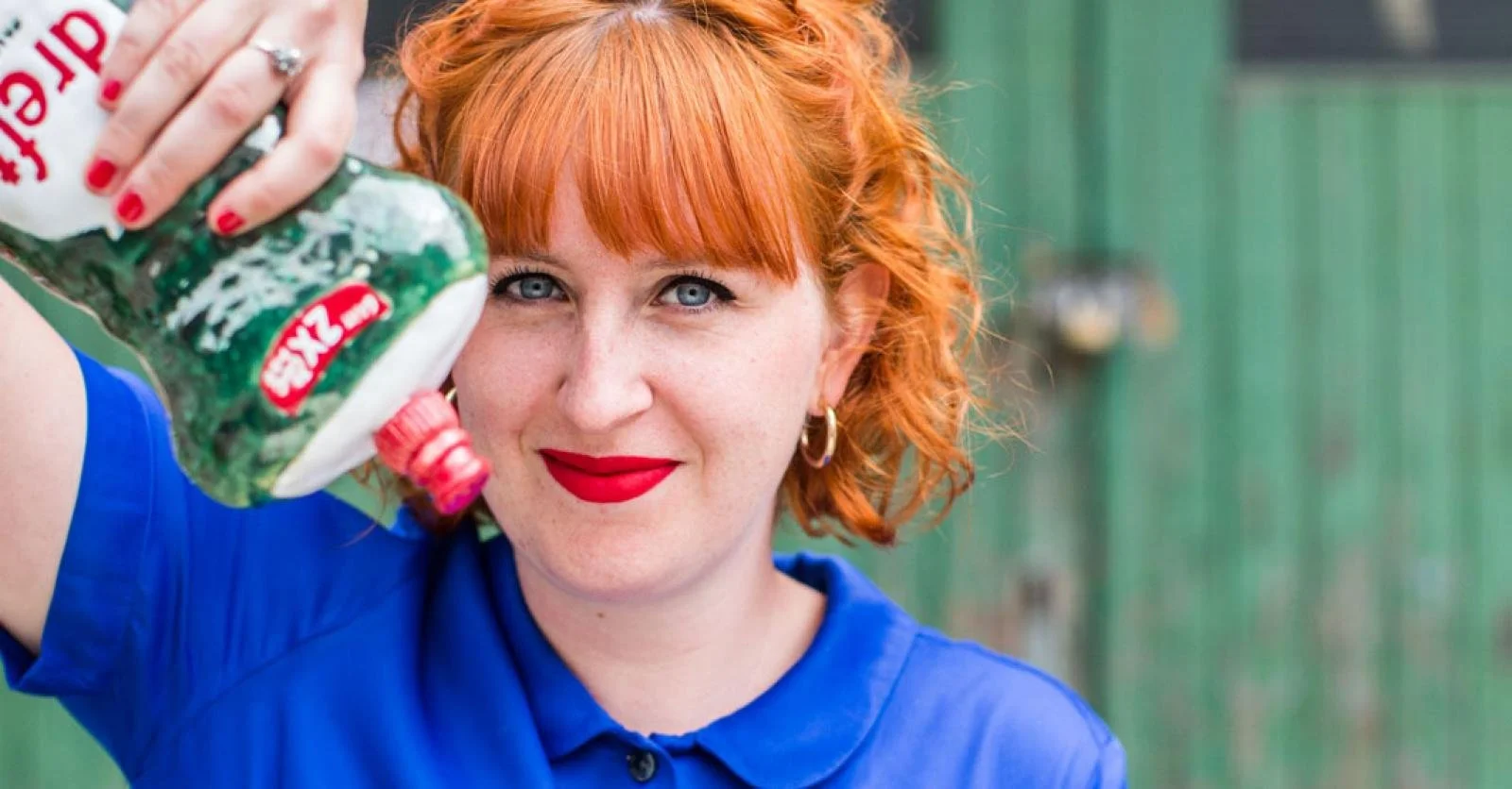 A woman with red hair and red lipstick holding a green soda bottle upside down near her face, smiling at the camera, wearing a blue shirt and hoop earrings, outdoors with a green background.
