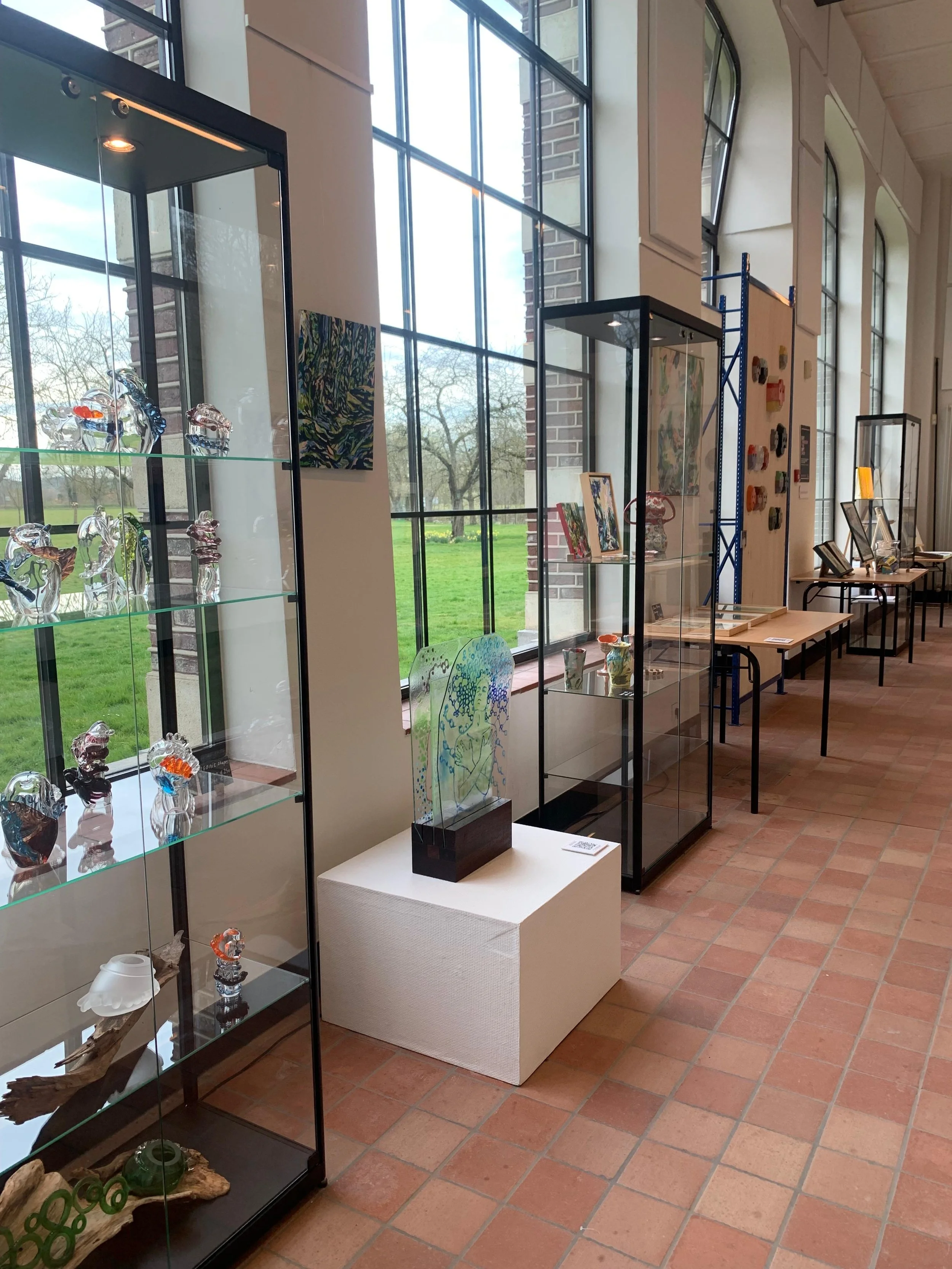 An art gallery with glass display cases holding glass sculptures and artwork. Large windows let in natural light, revealing a grassy outdoor area with trees.