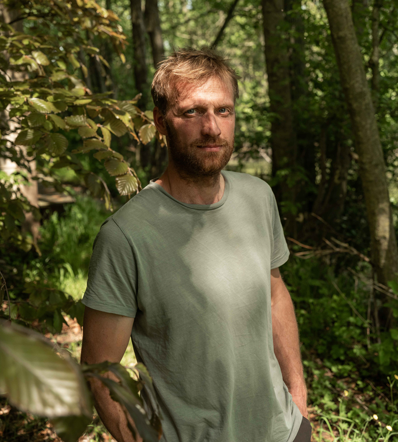 A man with light brown hair, beard, and mustache standing in a forest with sunlight filtering through the trees.