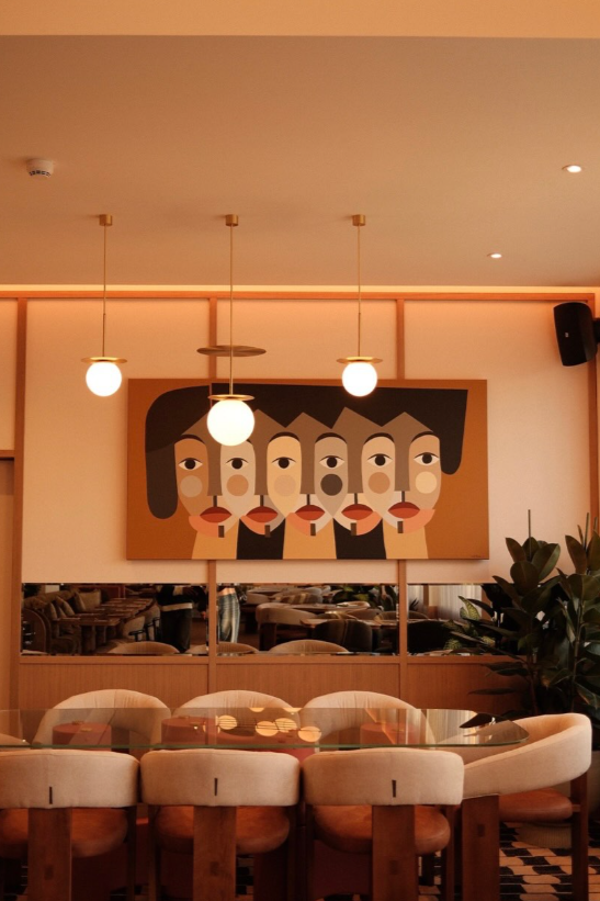 Interior of a restaurant or cafe with a large abstract art painting of five faces on the wall. There are three hanging light fixtures and beige upholstered chairs around a glass table.