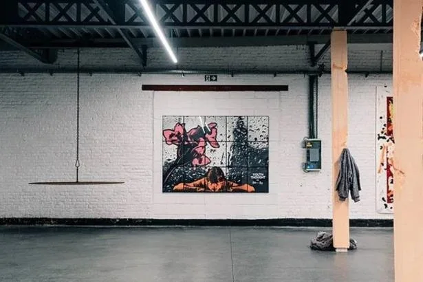 Binnenruimte met een witte bakstenen muur, een kunstwerk met bloemen en abstracte vormen, een lamp aan het plafond, een elektrische kast en een jas aan een houten pilaar.