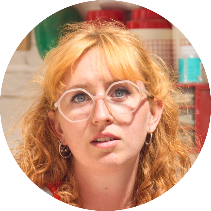A woman with curly red hair, wearing large clear glasses and hoop earrings, looking upward with a surprised or curious expression. The background includes various colorful bottles and containers.