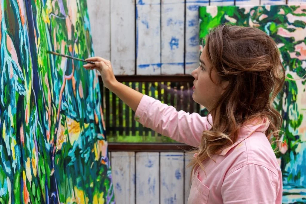 A woman with curly hair painting a colorful abstract mural on a wall outdoors.