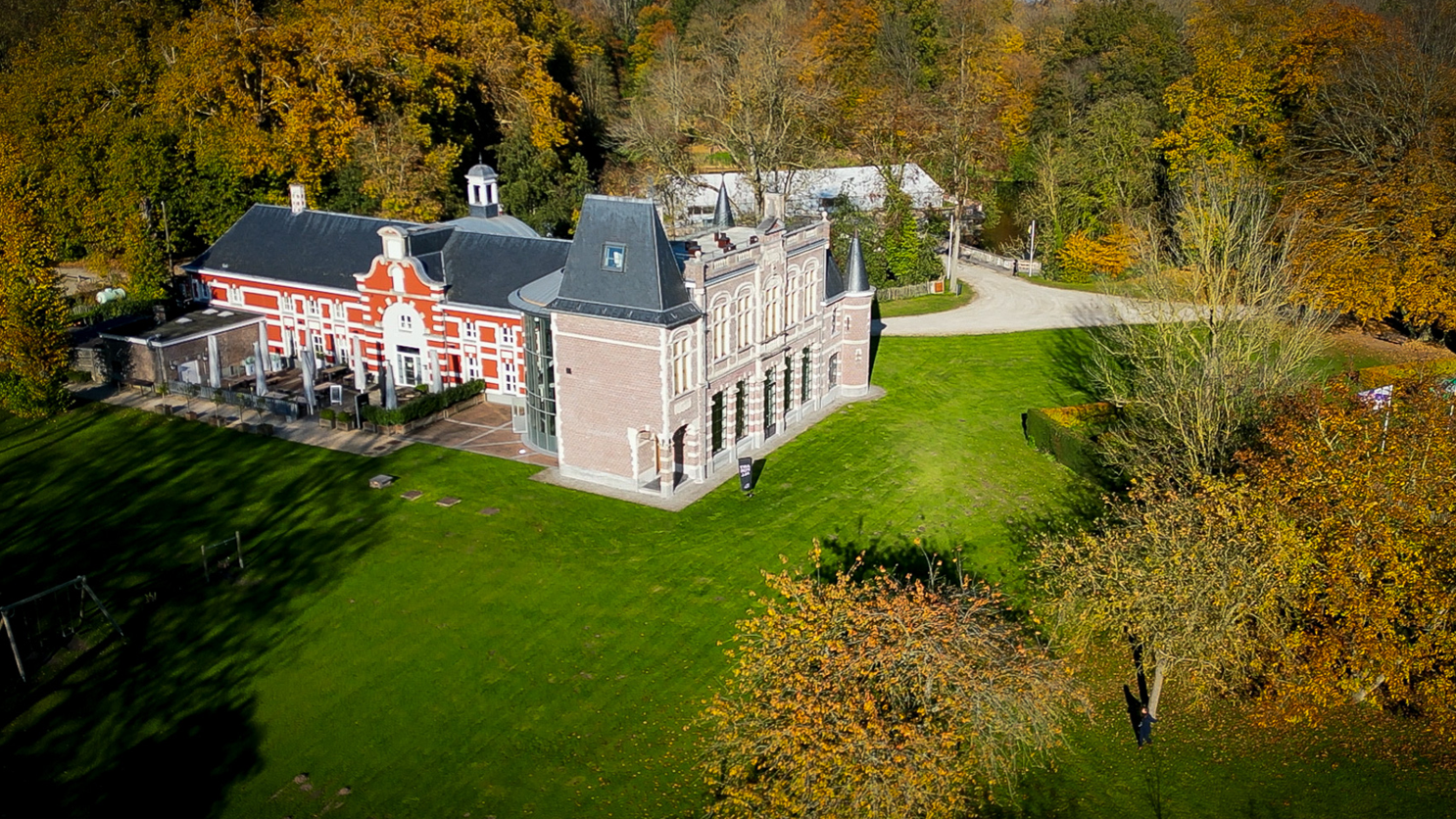 Aerial view of a historic building with red and white facade and gray roof, surrounded by green grass and trees with fall foliage.