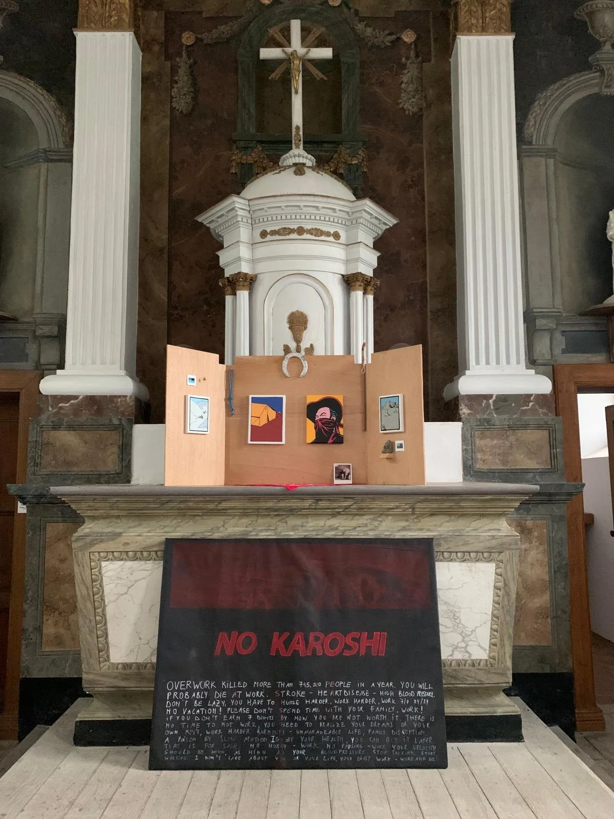 Inside a church, an altar with religious statues and artwork, including a prominent white crucifix and an ornate white structure, with a display of colorful paintings and a blackboard sign with the message 'NO KAROSHI' and text warning about overwork and health risks.