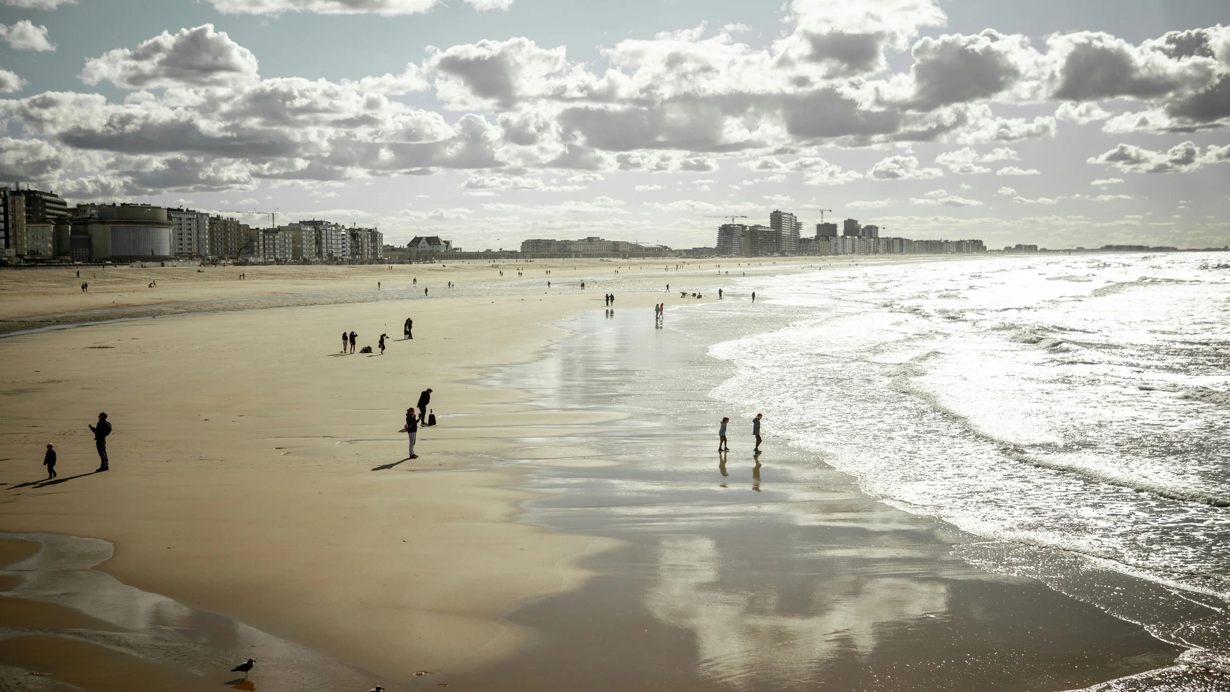 A beach scene with sandy shore, people walking and playing near the water, overcast sky with scattered clouds, and buildings along the coastline in the background.
