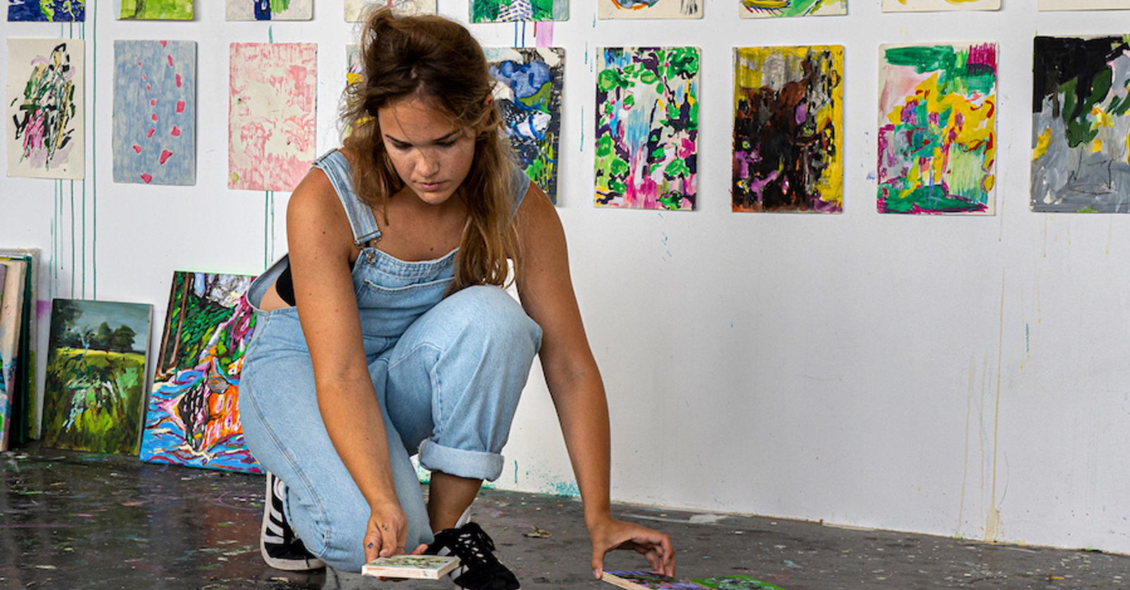 A young woman in overalls bends down in an art gallery, arranging her artwork on the floor. The gallery wall behind her displays colorful abstract paintings.