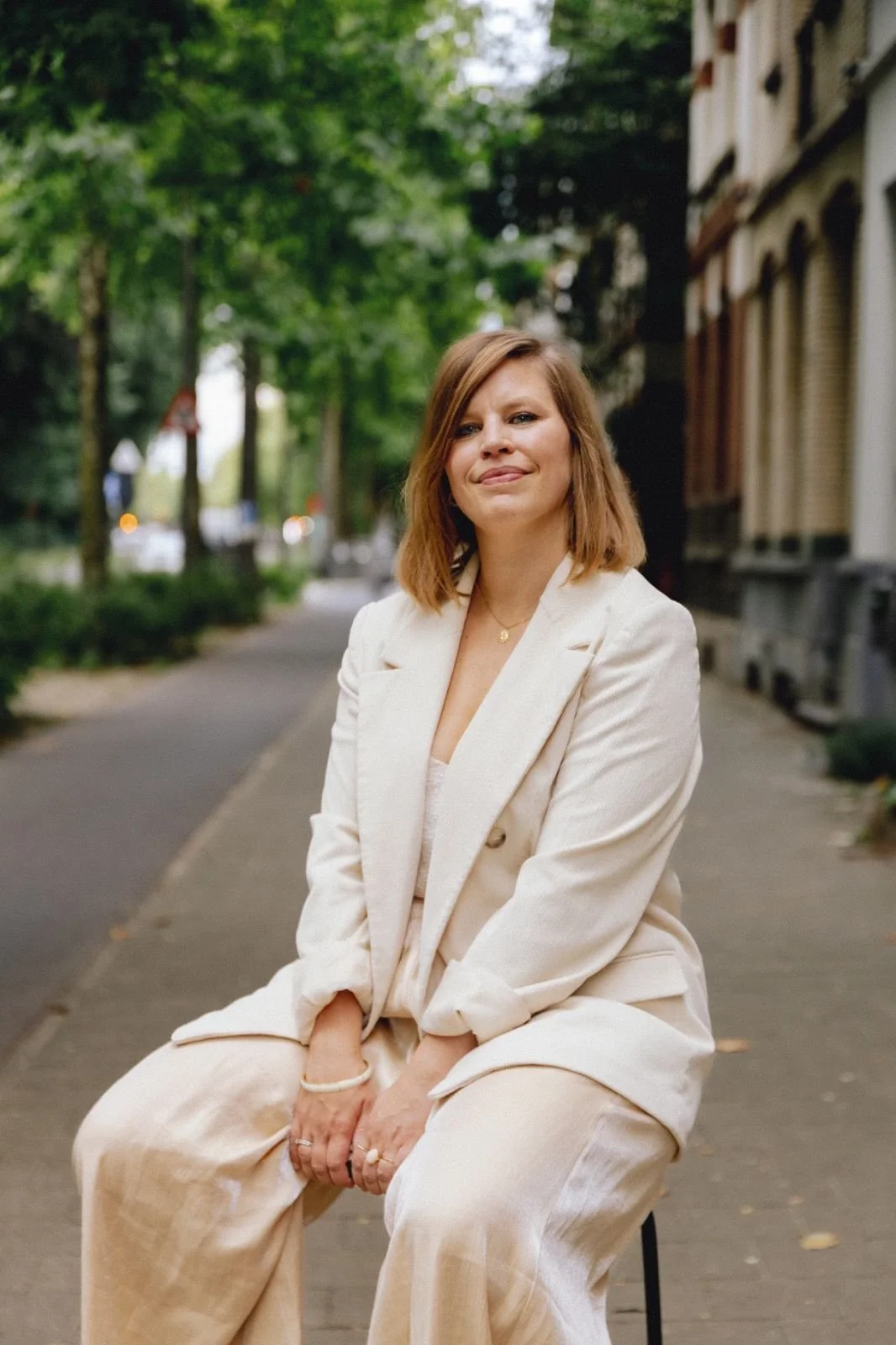 A woman with shoulder-length brown hair sitting on a stool on a sidewalk, wearing a cream-colored suit. She is outdoors with trees and buildings in the background.