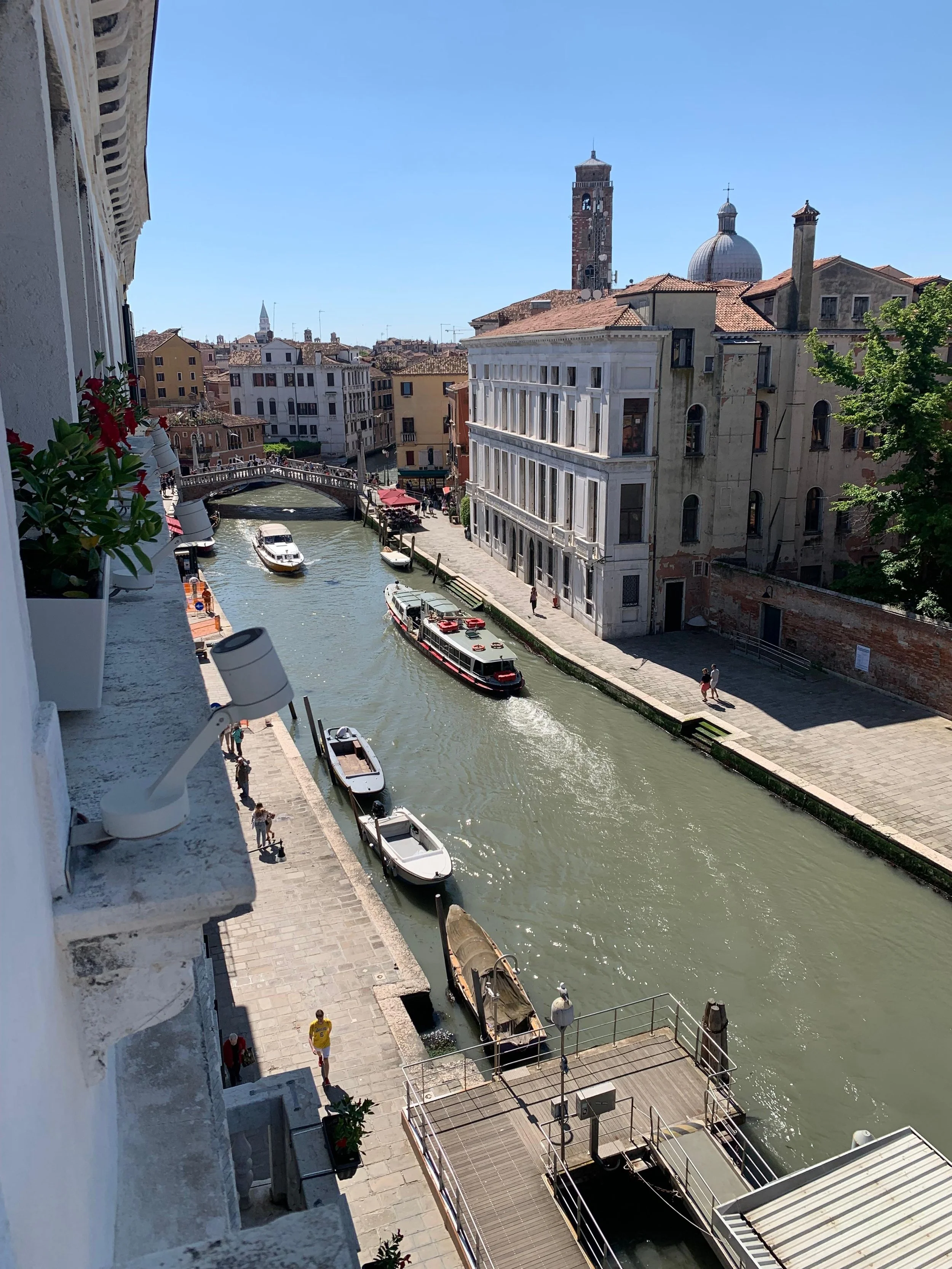 View of a canal in Venice, Italy, with boats along the sides, a bridge in the background, and historic buildings lining the waterway under a clear blue sky.