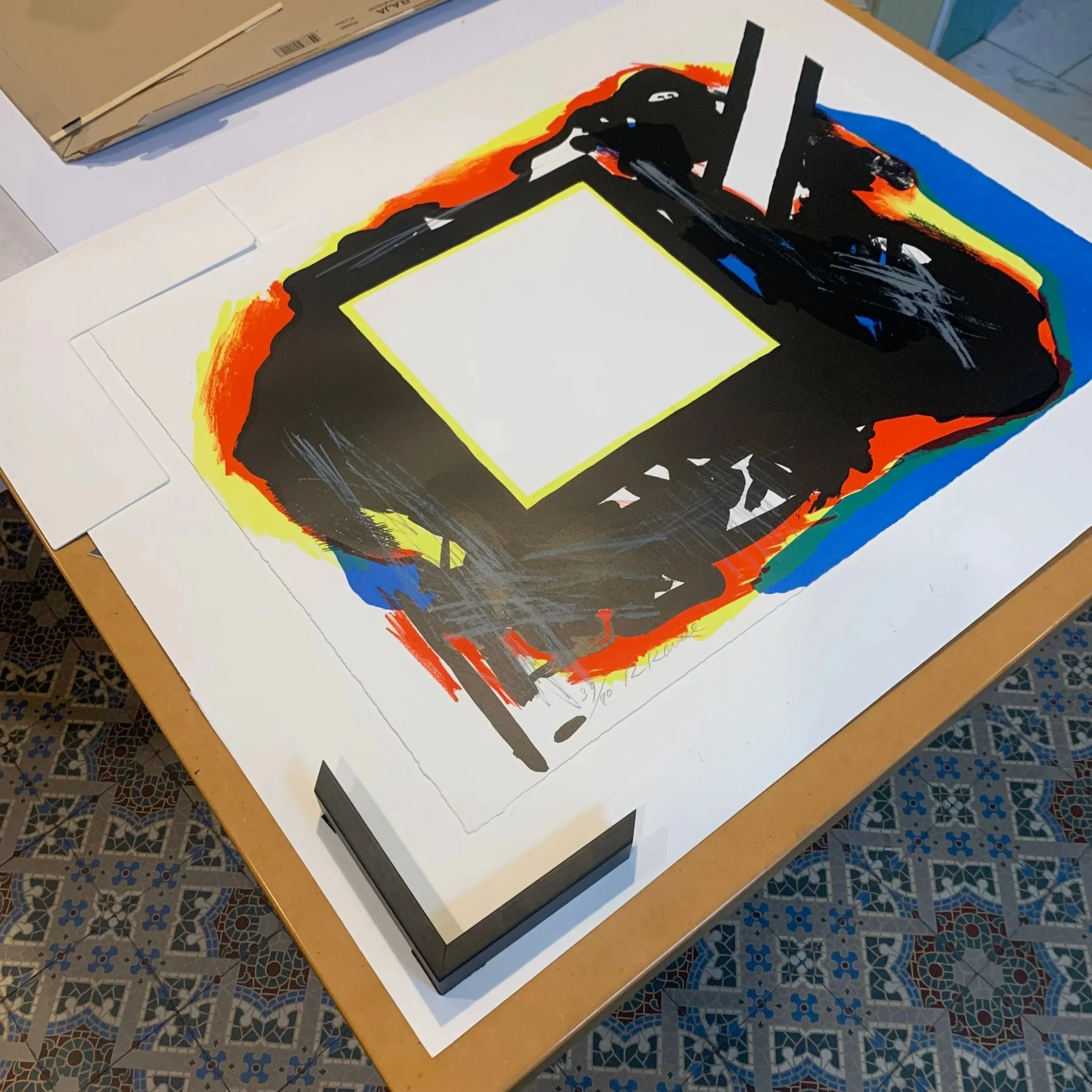 Abstract painting with bold black shapes, surrounded by red, yellow, blue, and green accents, on white paper, displayed on a wooden table.