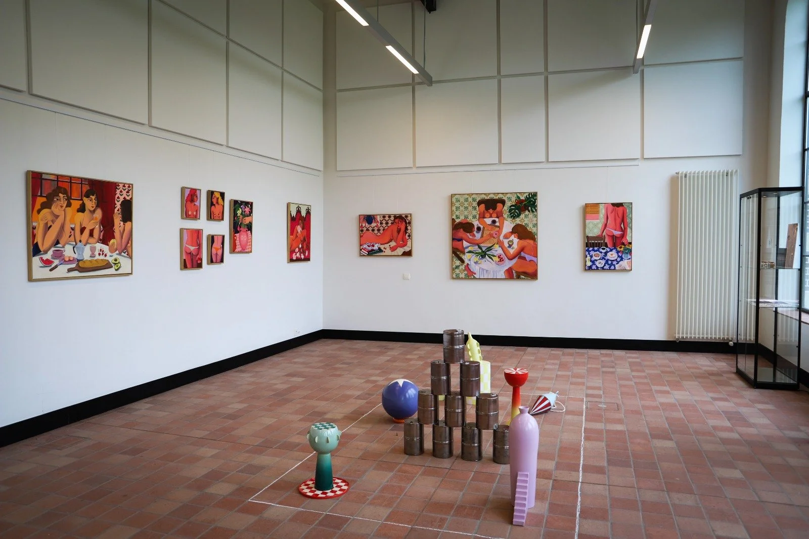 Art gallery with colorful paintings on white walls and a tiled floor, displaying various vibrant artworks, with sculptures and colorful objects on the floor in the center of the room.