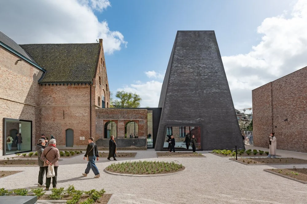 Een binnenplaats met oude bakstenen gebouwen en een nieuw modern zwart betonnen gebouw in het midden, met mensen die rondlopen en foto's maken.