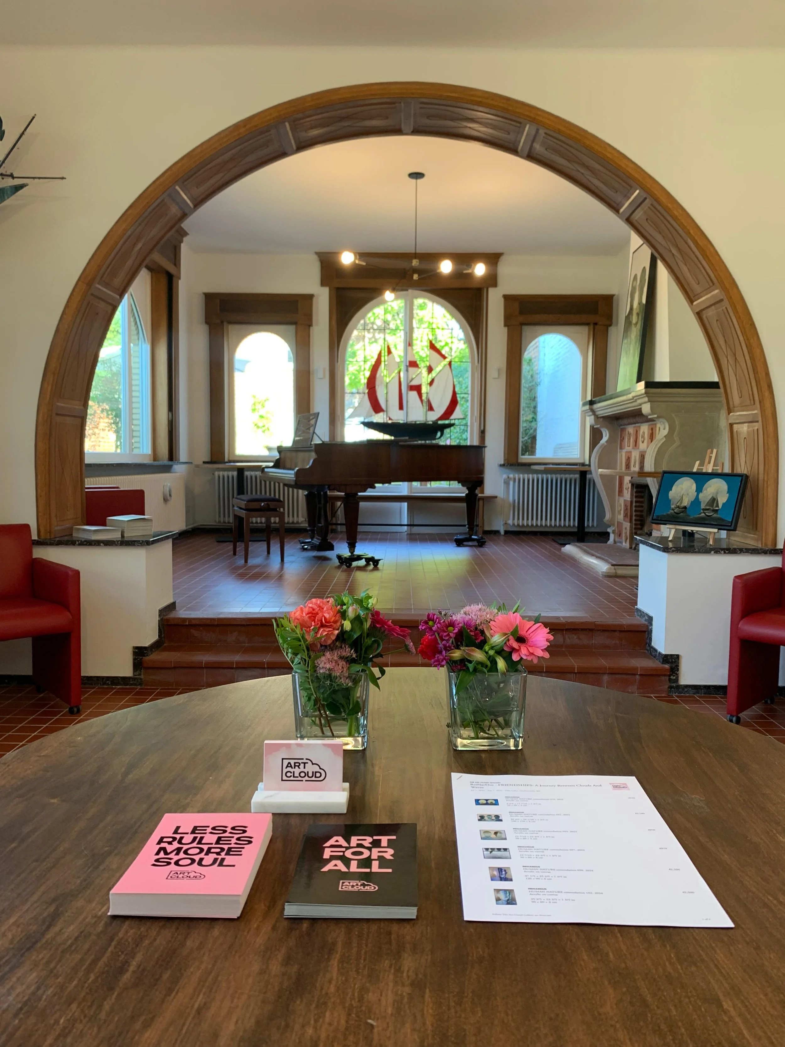 Interior of a room with a grand piano on a small stage, decorated with a model ship, and a large stained glass window with greenery outside. A round table in the foreground has books titled "Less Rules More Soul" and "Art For All," two floral arrangements, and a brochure or program.