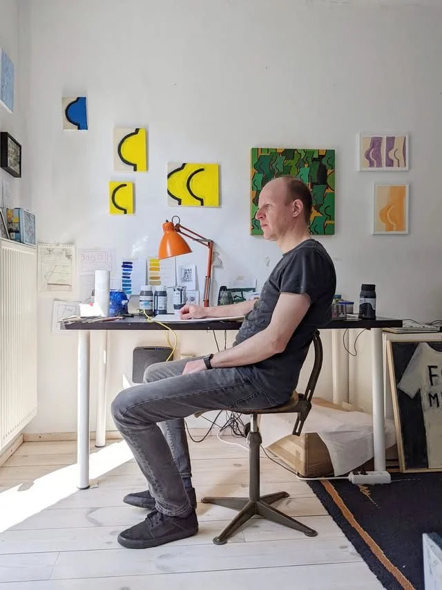 A man sitting at a desk in a room with colorful abstract wall art, with art supplies and a lamp on the desk.