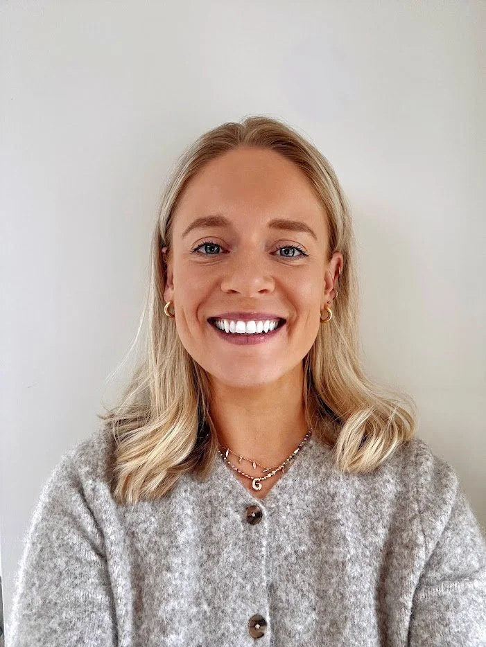 A smiling woman with blonde hair, blue eyes, wearing gold hoop earrings, layered necklaces, and a gray button-up sweater standing against a plain white background.