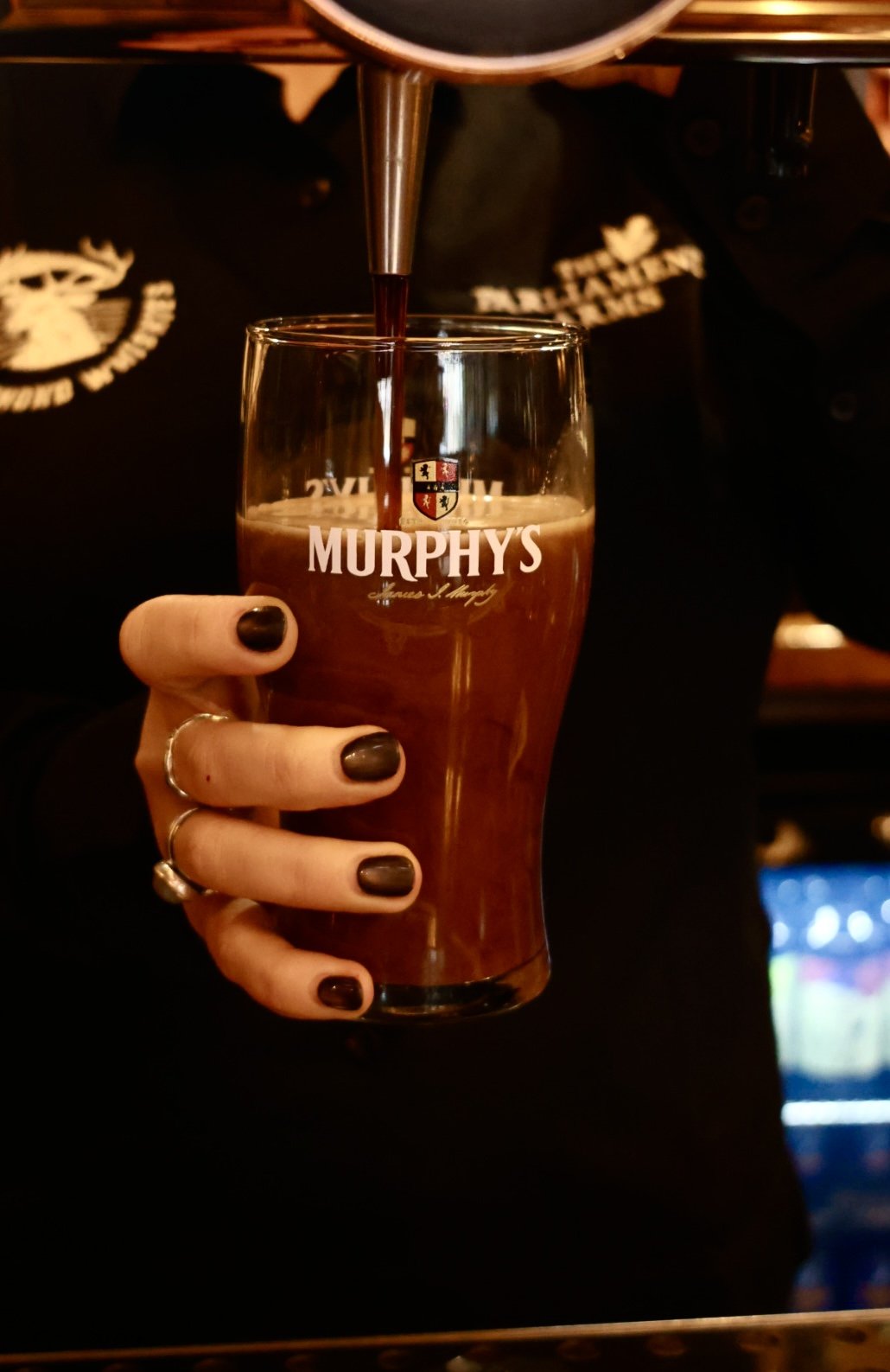 A tall glass of Amstel Bier beer with a foamy head on a wooden table, with a glass of white wine, a bowl of food, and a salad in the background in a restaurant setting.