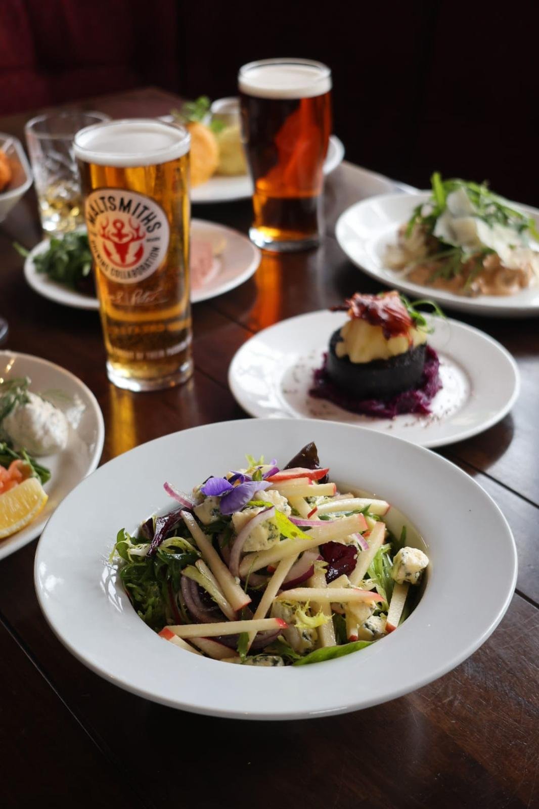 A table set with various dishes, including a colorful salad with edible flowers in the foreground, two glasses of beer, and other plated entrees in the background.