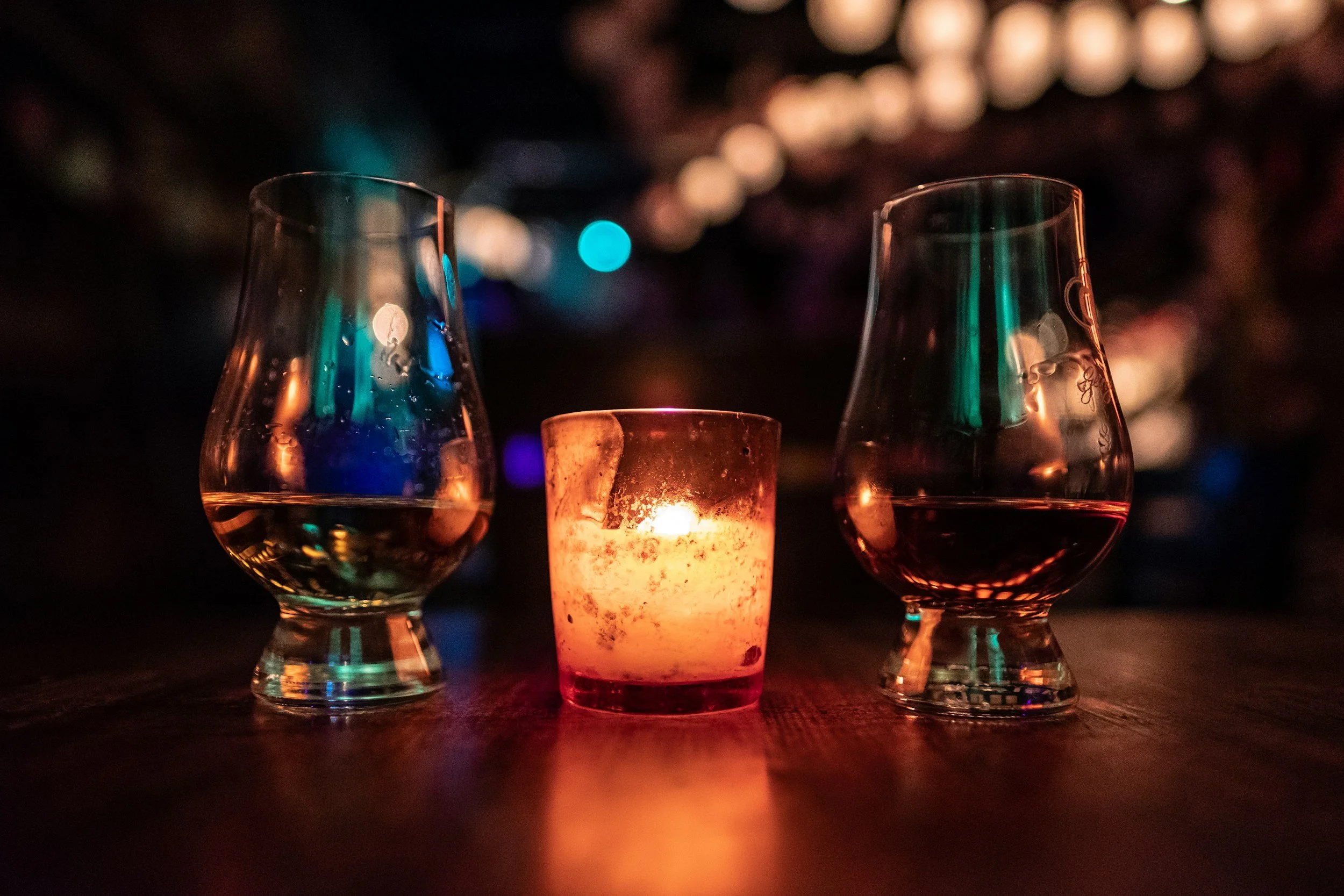 Two glencairn glasses with whiskey and a lit candle in the center, on a wooden surface, with blurred colorful lights in the background.
