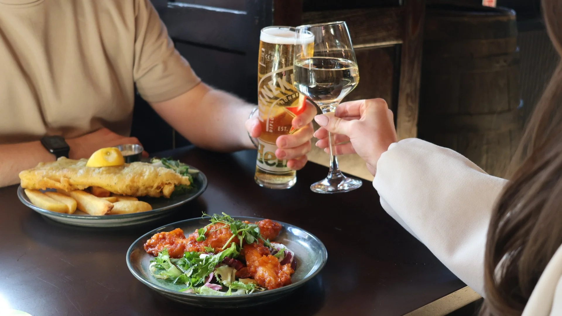 Close-up of a plated meal featuring a pork chop topped with shrimp in sauce, a side salad with cherry tomatoes, and a portion of potato chips in the background, with a glass of red wine above.