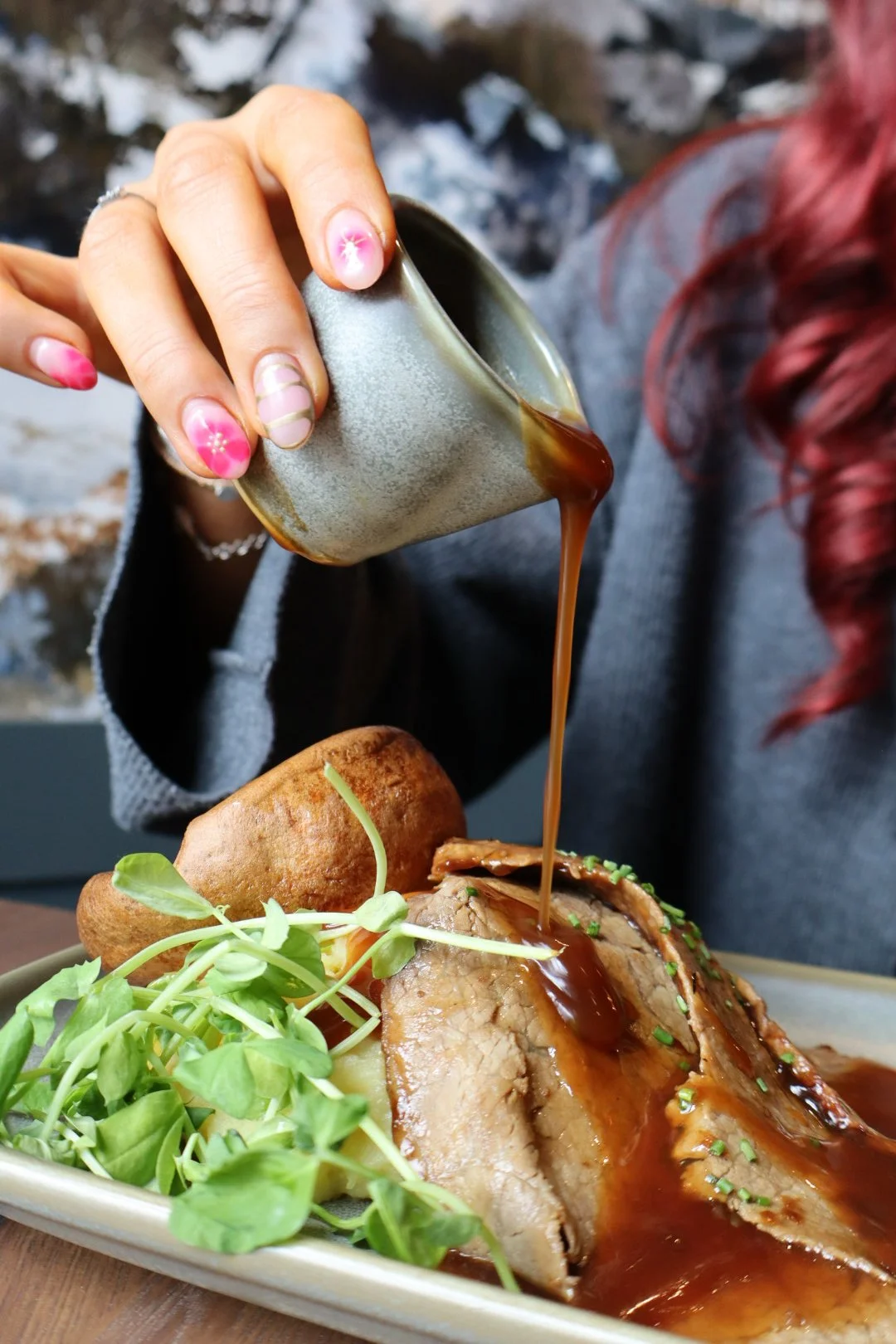 Person pouring gravy over a roasted meat dish topped with chopped herbs, with a side of potatoes and greens, and a person with red hair wearing a gray sweater.