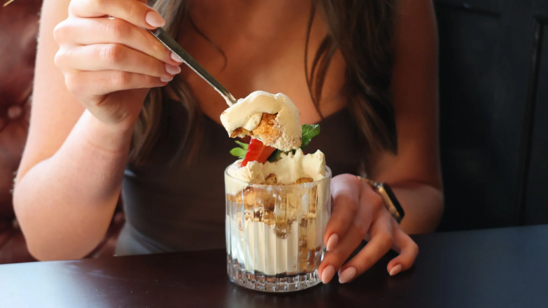 A slice of cheesecake garnished with blackberries, a mint leaf, and powdered sugar on a white plate, with a glass of parfait dessert in the background on a white plate.