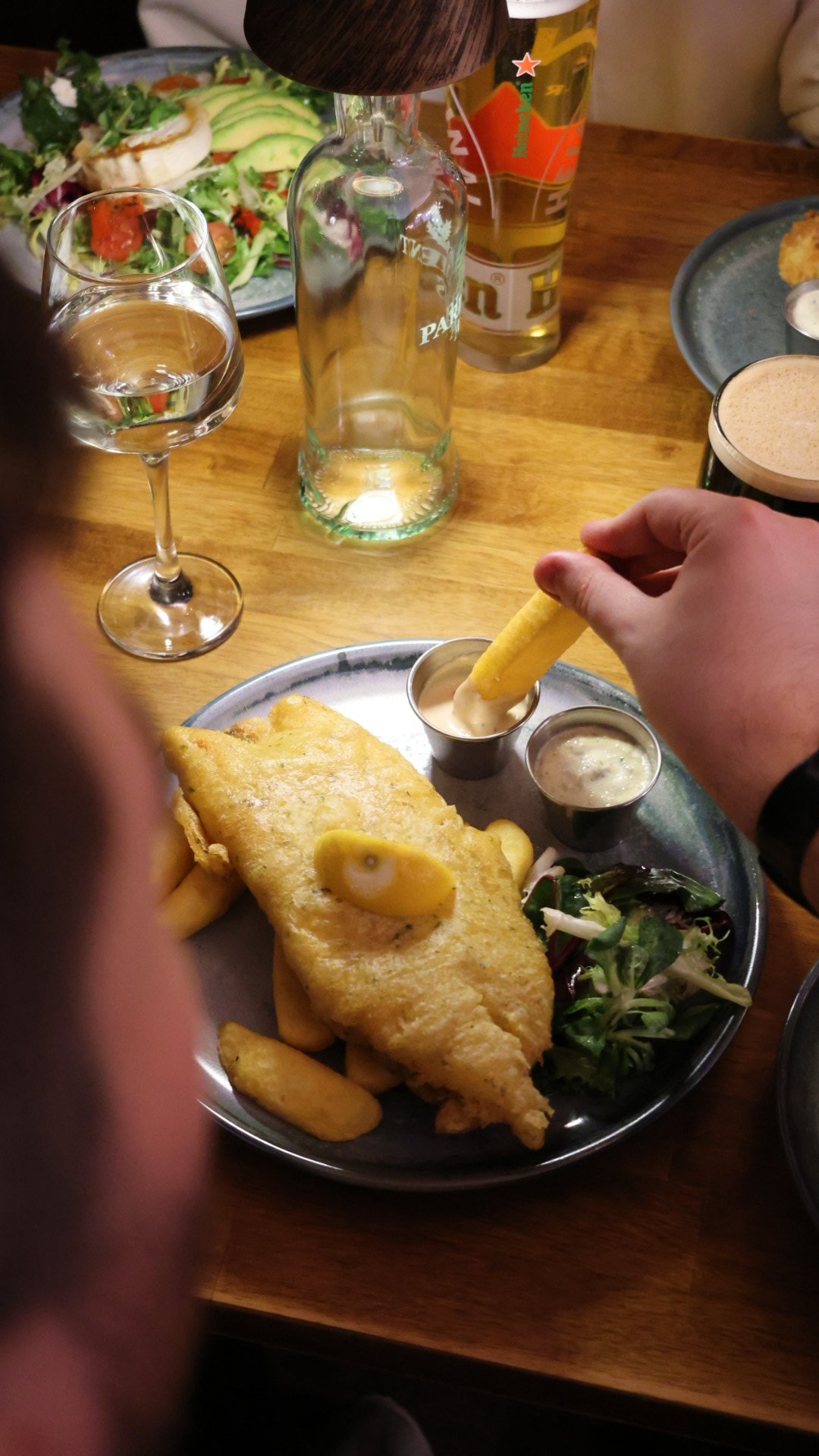 A hand squeezing lemon over a plate of fish and chips with green peas and tartar sauce, surrounded by glasses of beer in a pub setting.