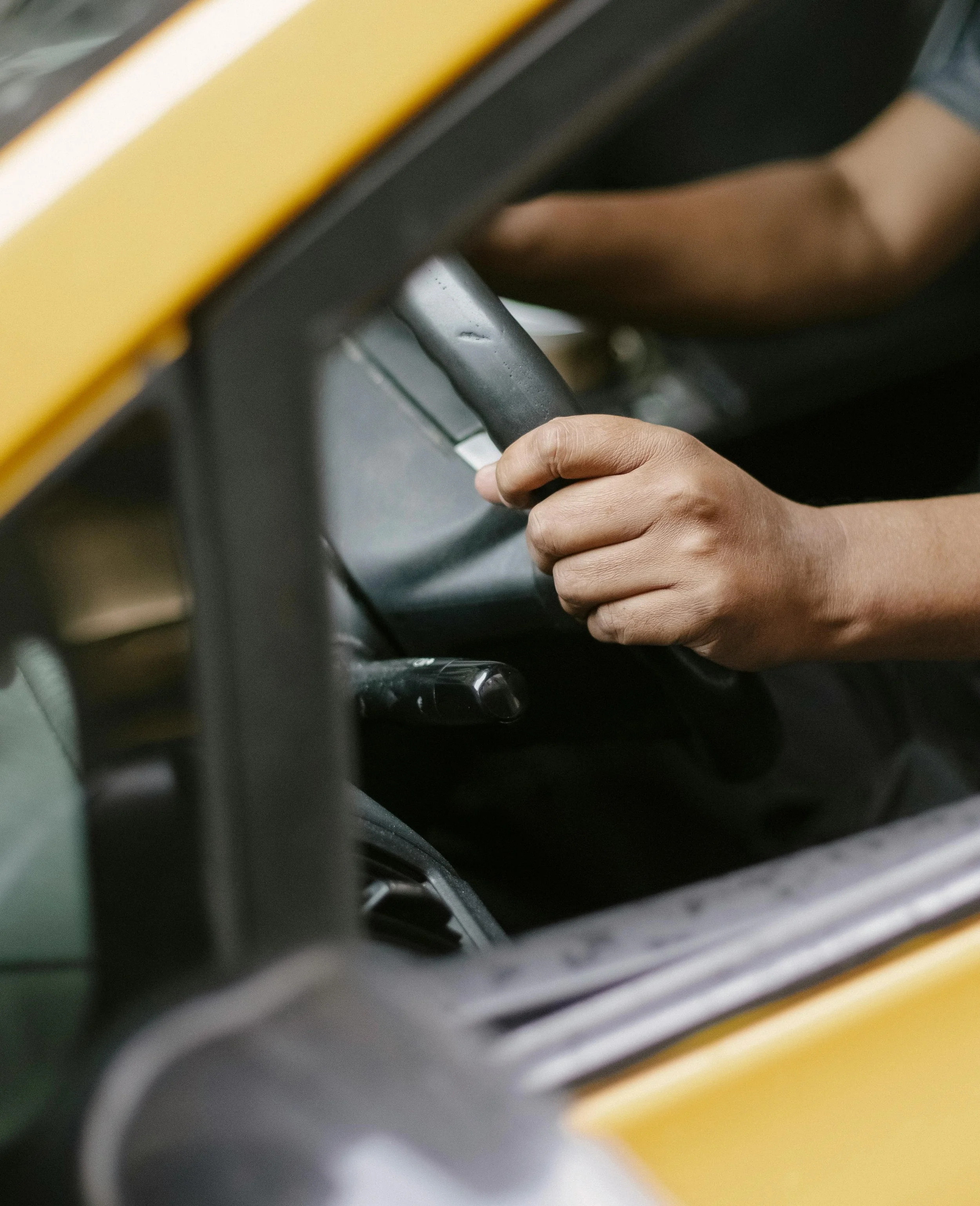 Nahaufnahme einer Hand, die das Lenkrad eines Fahrzeugs greift, vermutlich eines Baustellenfahrzeugs, mit gelbem und schwarzem Fahrzeugsegment sichtbar.