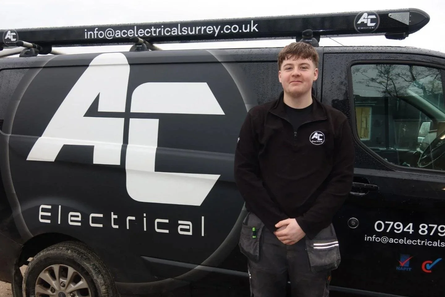 Young man standing in front of a black van with the URL 'urrey.co.uk' on it, wearing a black AC Electric jacket, with raindrops on the vehicle.