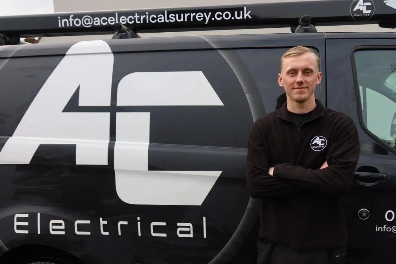 A young man with blond hair smiling, wearing a black AC Electric jacket, standing in front of a black van with AC Electric branding.