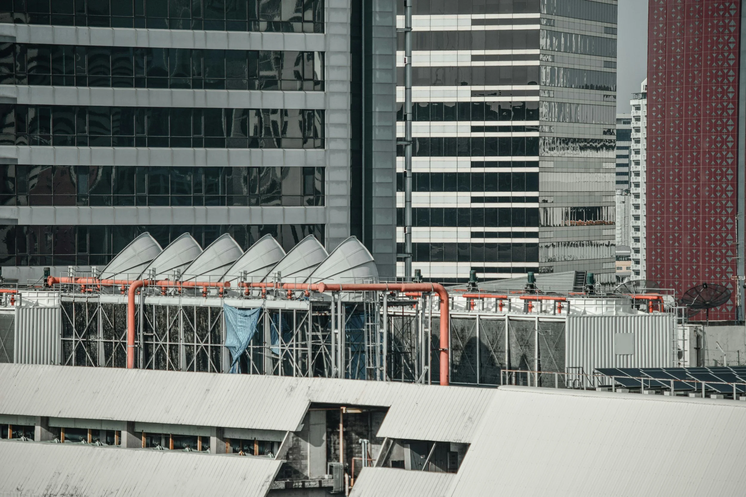 Close-up view of a cityscape showing modern high-rise buildings with glass facades and a rooftop containing piping, scaffolding, and a satellite dish.