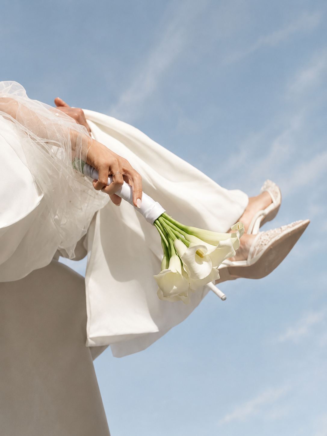 Braut in weissem Kleid hält einen Strauss weisser Calla-Lilien vor blauem Himmel während dem Brautpaarshooting. Bild KI generiert.