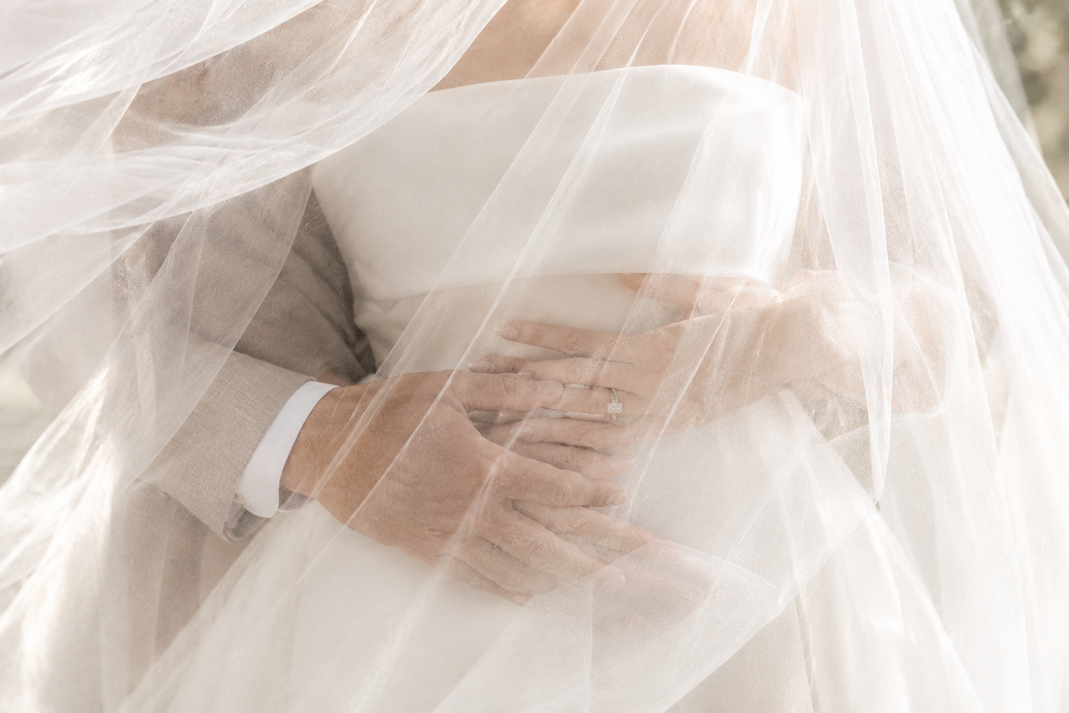 Nahaufnahme eines Brautpaares unter einem transparenten weissen Schleier, mit ineinandergelegten Händen und sichtbarem Ehering. Bild KI generiert.