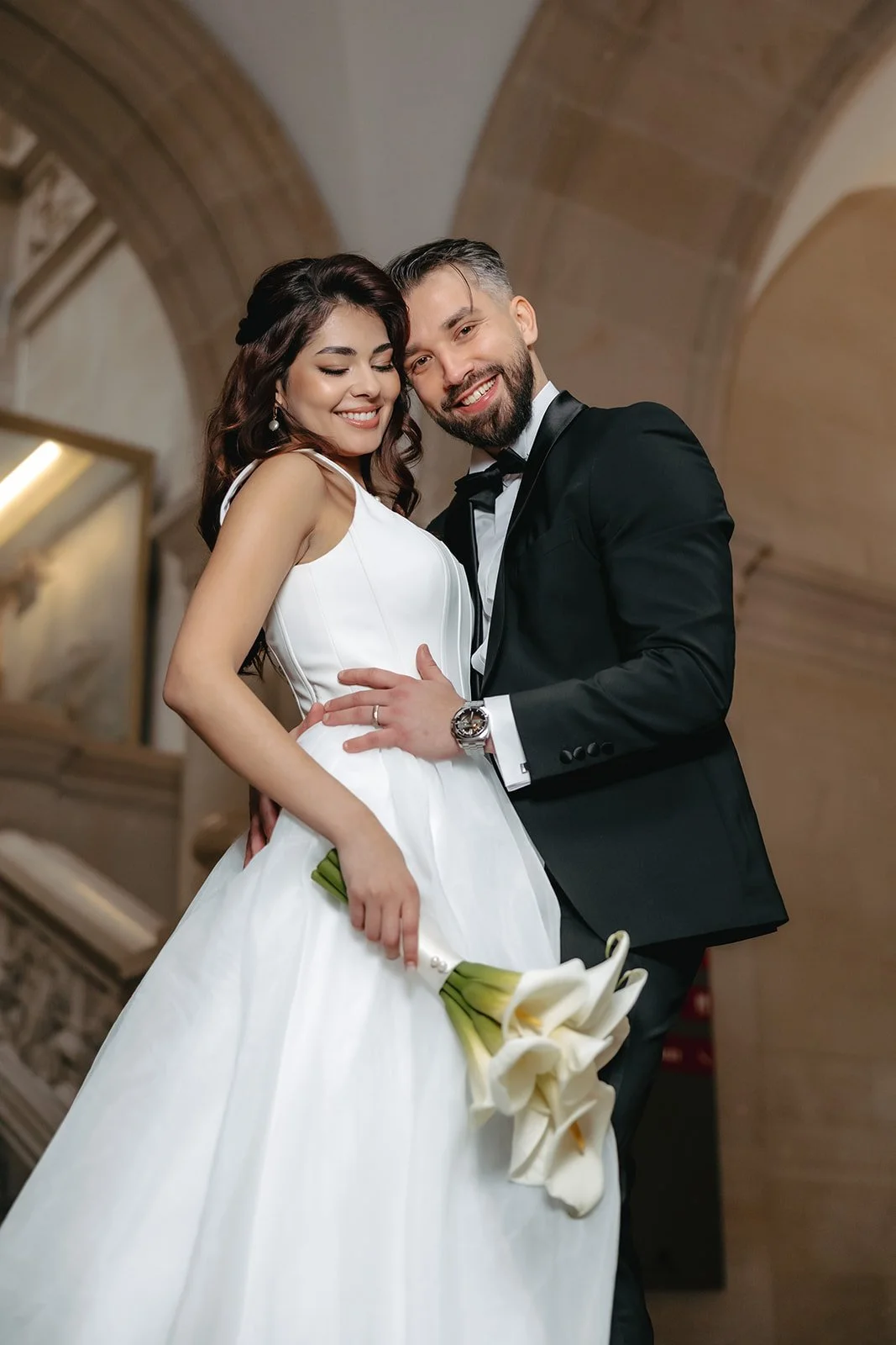 Lächelndes Brautpaar in weissem Hochzeitskleid und schwarzem Smoking mit weissen Calla-Lilien in einer romantischen Hochzeitslocation. Bild von Gloria Velvet.