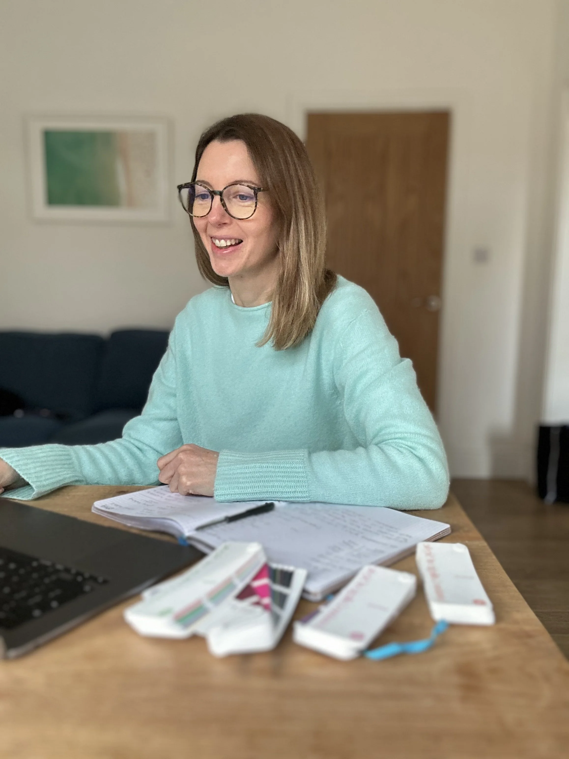 A woman with shoulder-length brown hair, glasses, and a mint green sweater, smiling while sitting at a wooden table with a laptop, an open notebook, and medical supplies such as test strips.