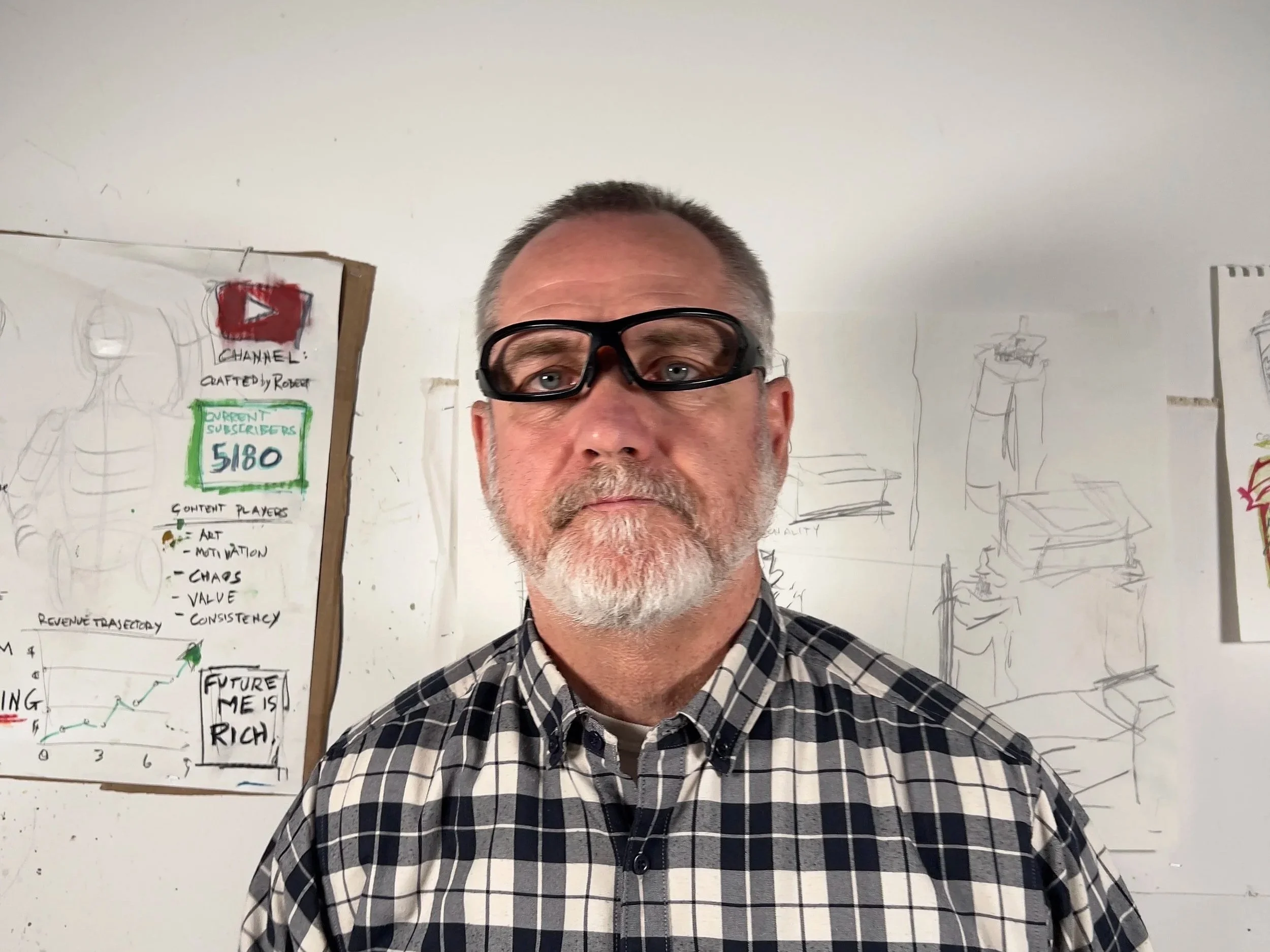 A middle-aged man with short gray hair and a beard wearing glasses on his forehead and a black-and-white checkered shirt, standing in front of a whiteboard with diagrams and notes.