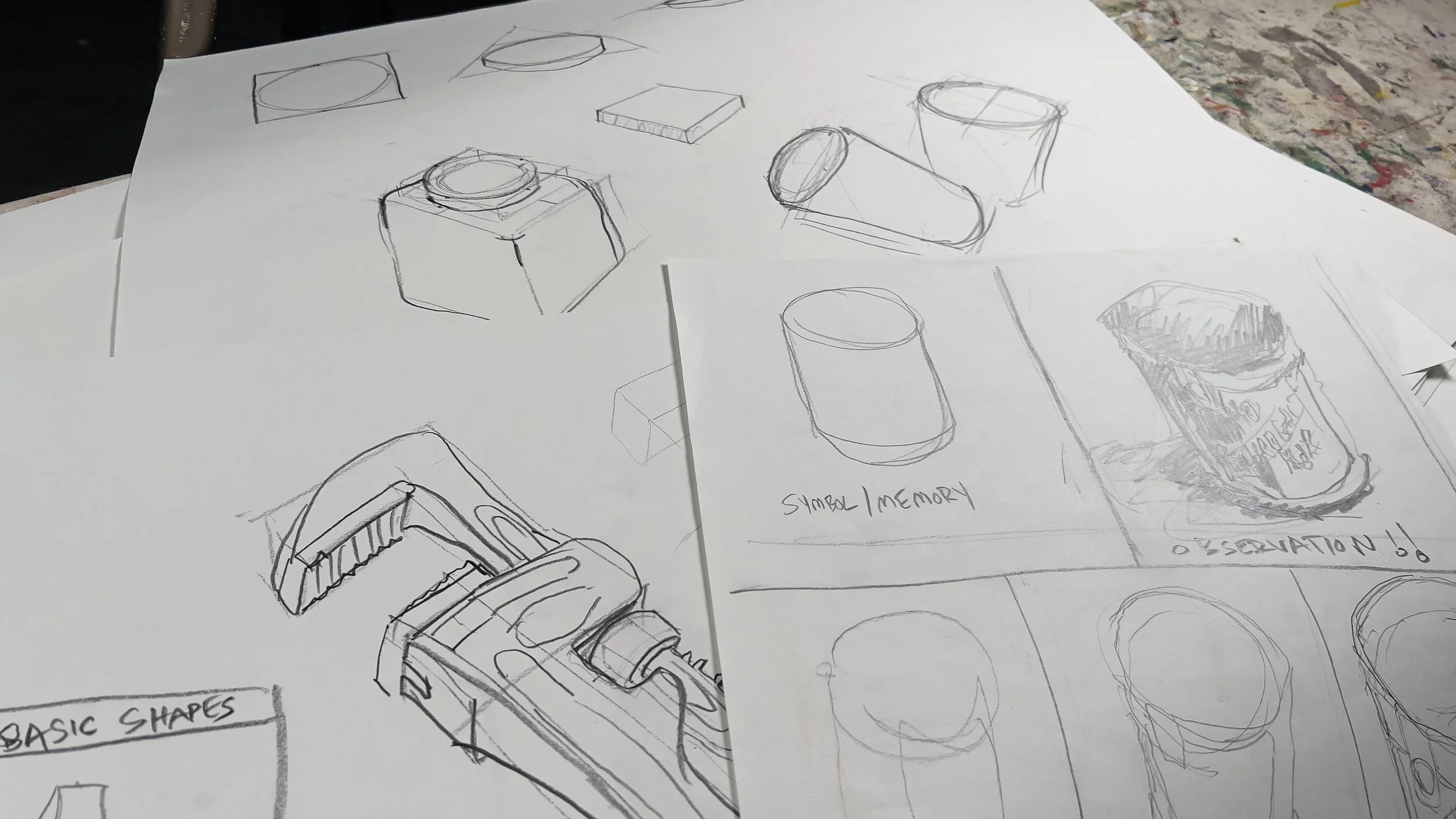 Sketches of various object designs, including a rectangular shape, a cylinder, a square, and a can labeled 'symbol/memory' and 'exploration', drawn on sheets of paper with some colorful paint splatters in the background.
