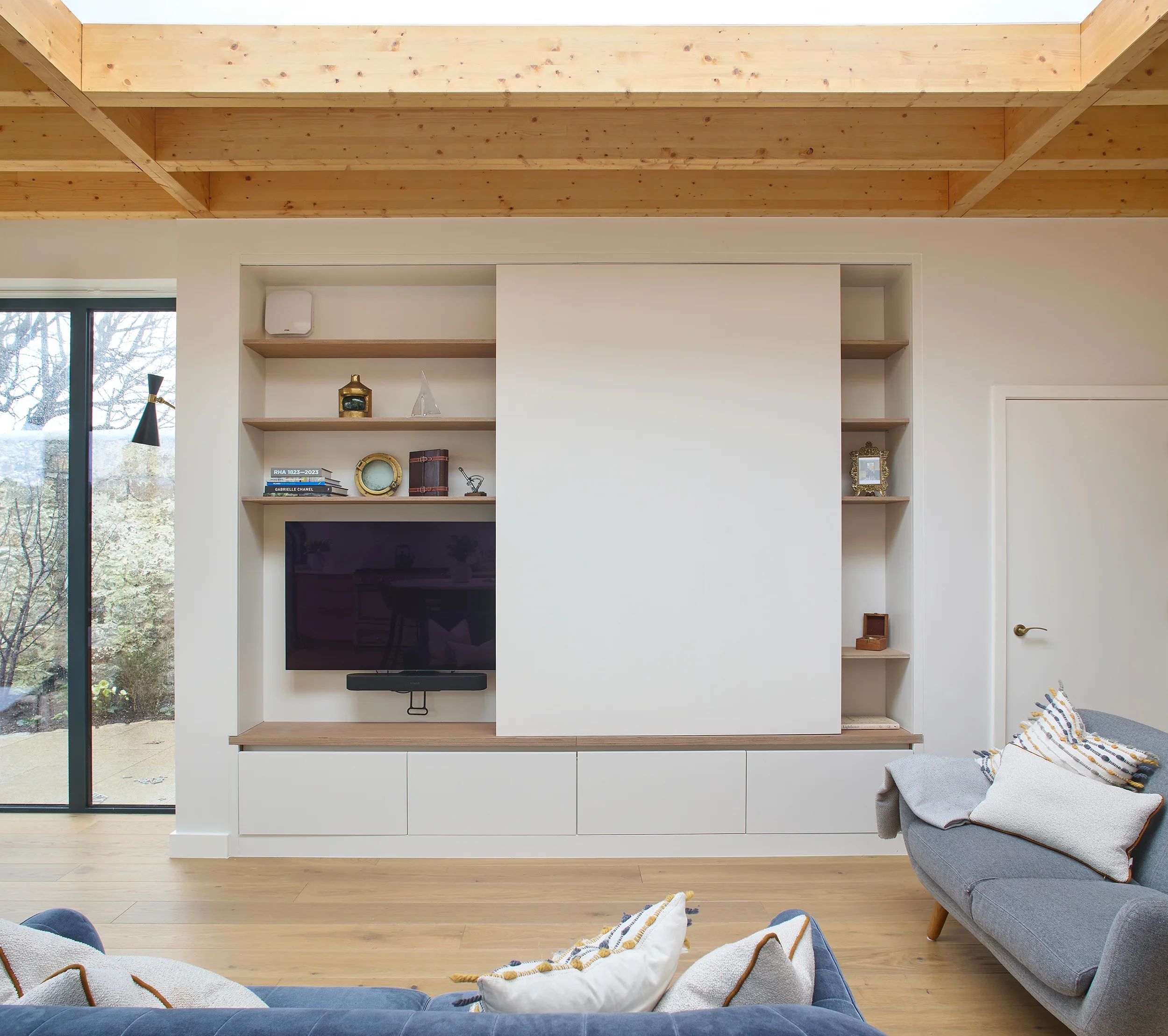 Modern living room with built-in white entertainment center, shelves with decorative items, flat-screen TV, and a gray sofa with cushions.