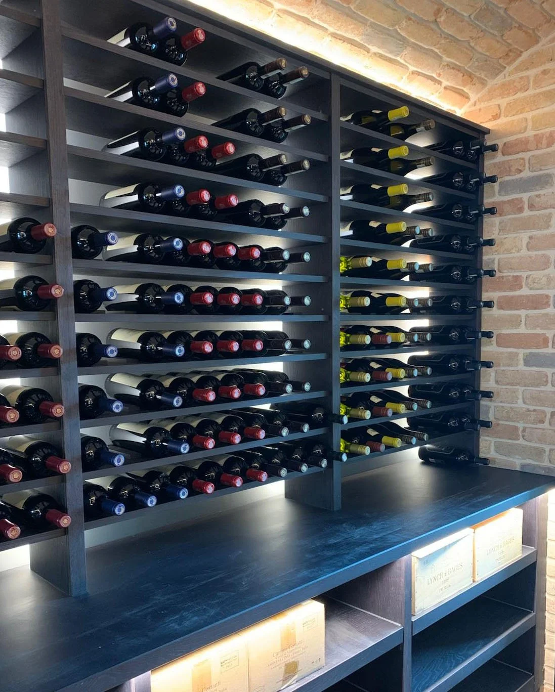 Wine bottles stored on black wooden racks against brick wall, with soft lighting.