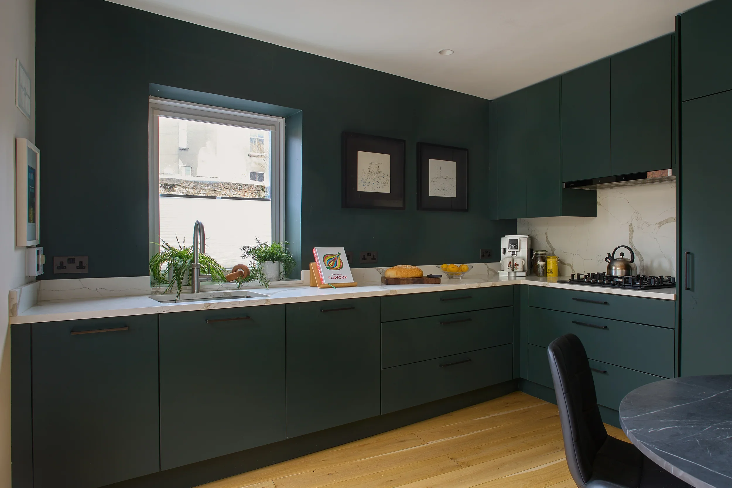 Modern kitchen with dark green cabinets, white marble countertops, a window above the sink, potted plants on the windowsill, a loaf of bread, a bowl of lemons, a coffee machine, a kettle, and framed artwork on the dark green wall.