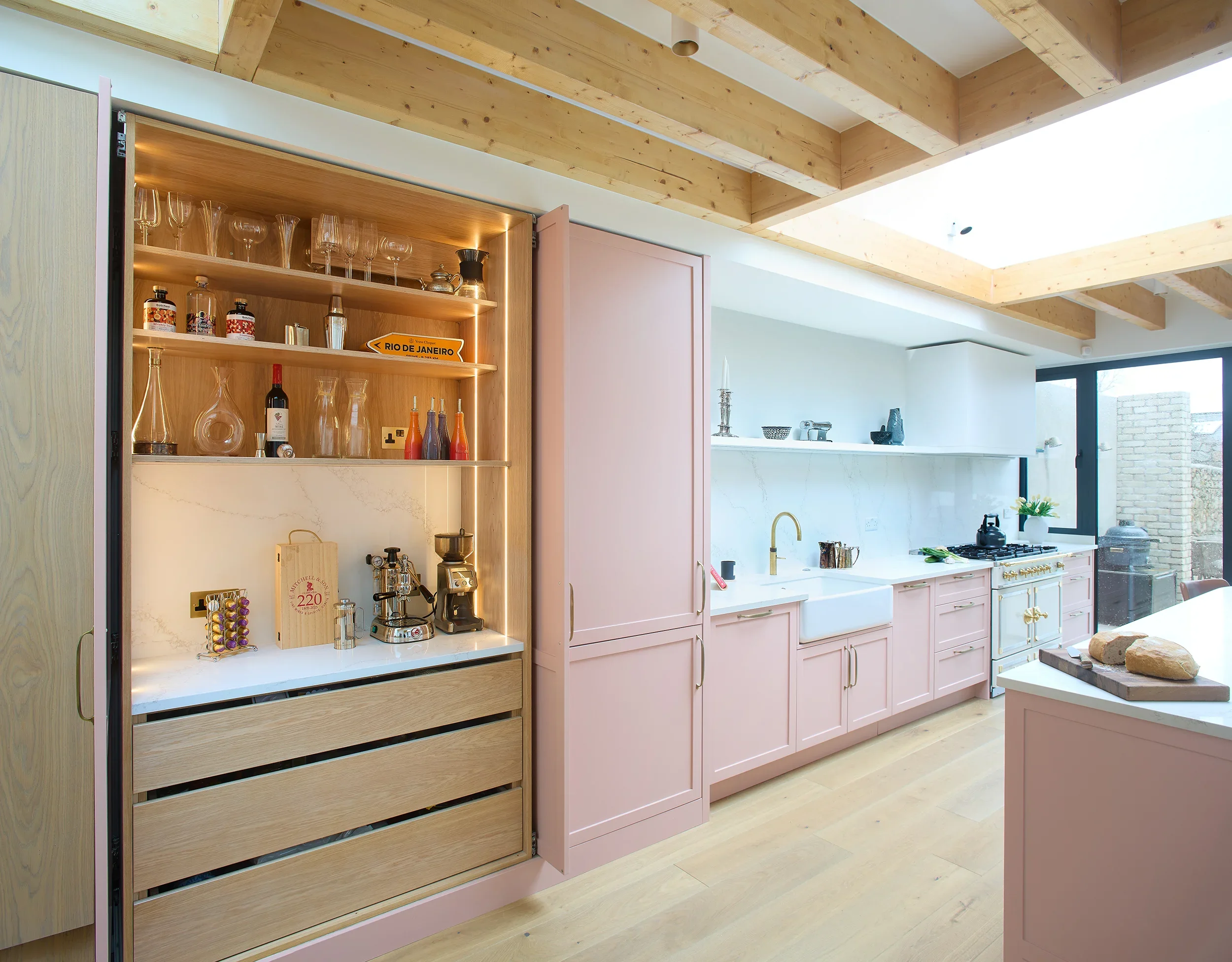 Blush pink shaker kitchen with large central island, exposed timber beam ceiling, white marble backsplash, and brass hardware in a modern open-plan home