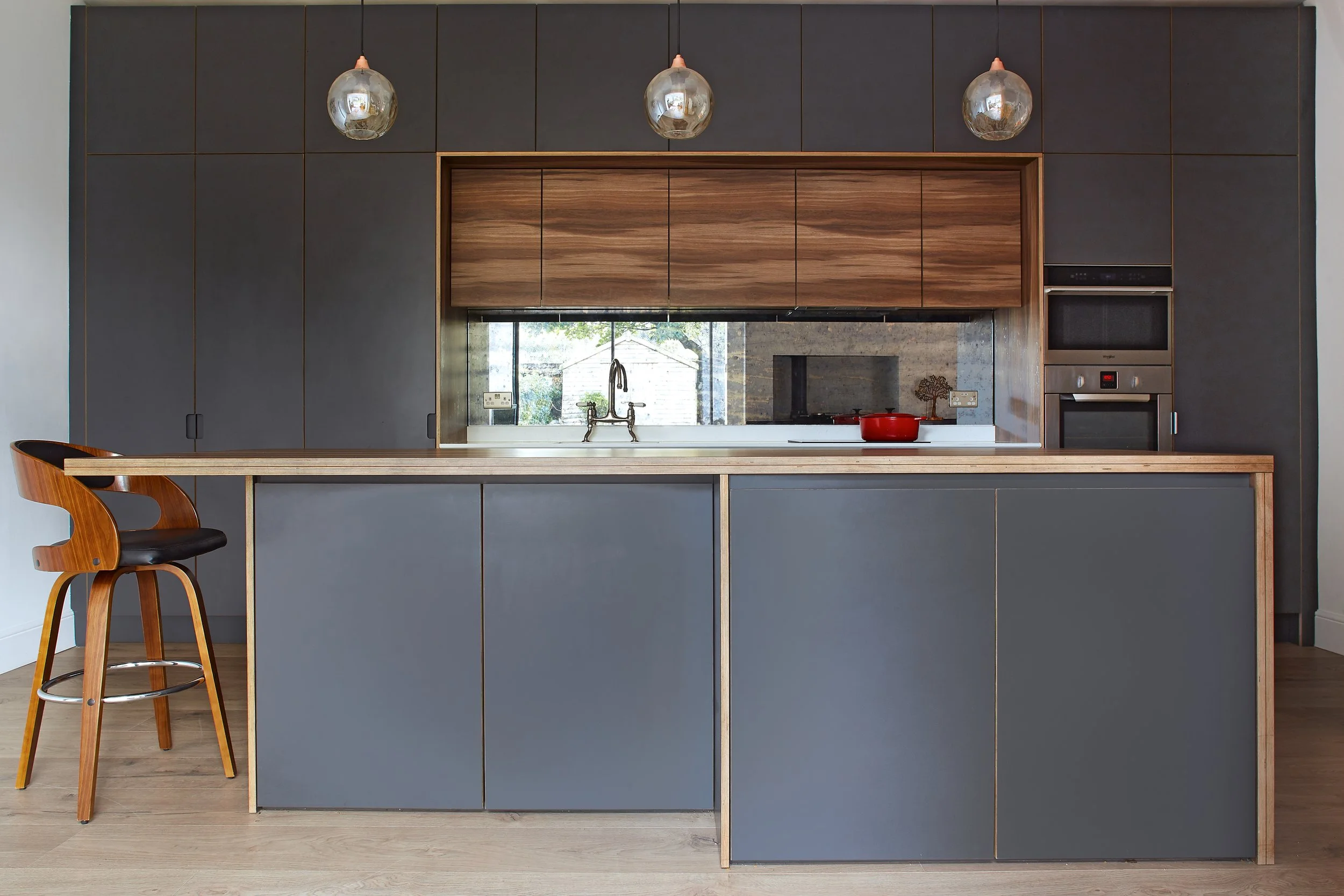Modern kitchen with gray and wood cabinets, a kitchen island with a wooden countertop, two hanging glass pendant lights, a window with a view outside, built-in oven and microwave, and a red pot on the stove.