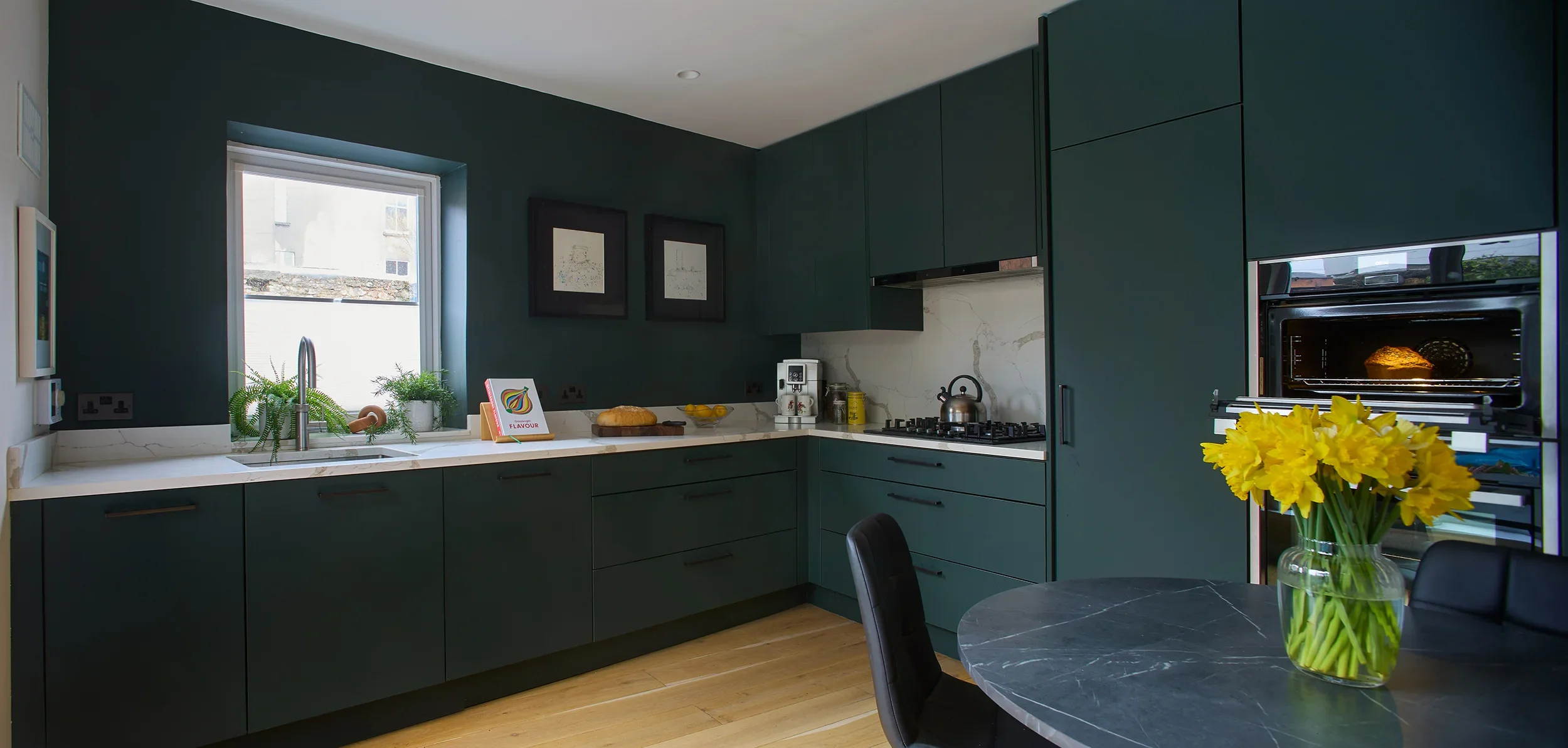 Modern kitchen with dark green cabinets, marble countertops, and a window above the sink. The kitchen features a gas stove, coffee machine, and a vase of yellow daffodils on a round black marble table with black chairs.