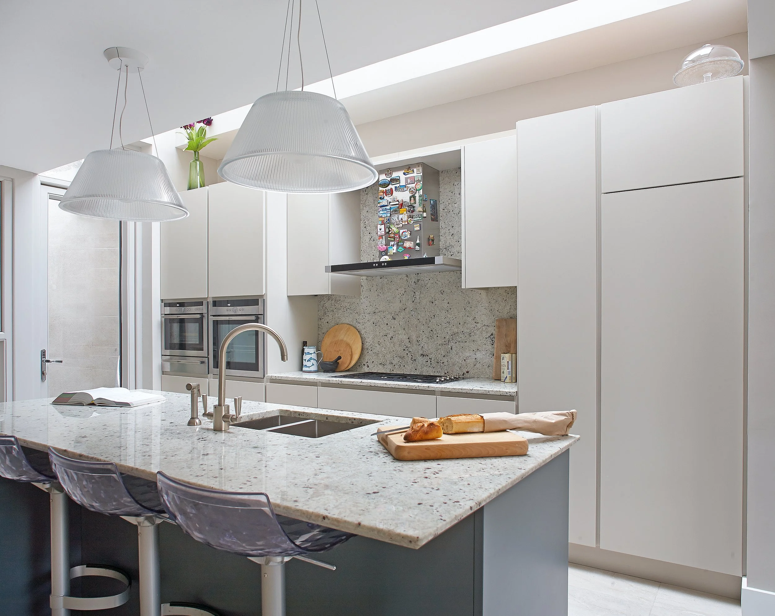 Modern white kitchen with granite island, pendant lighting, and built-in appliances, with a cutting board of bread on the counter.