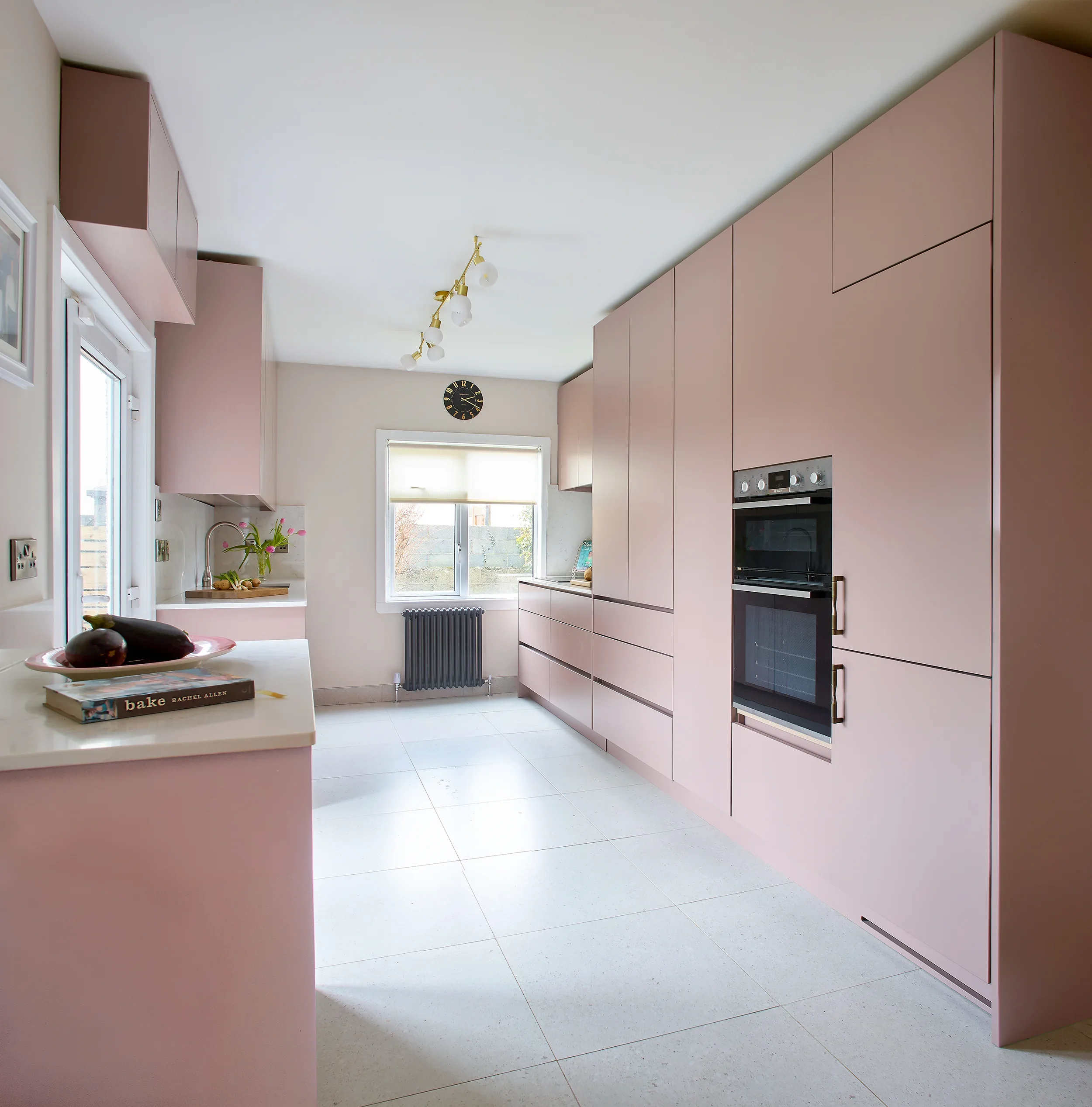 A modern kitchen with pink cabinets, a white countertop, a window, a black radiator, and a wall clock, with a book titled 'Bake' on the counter.