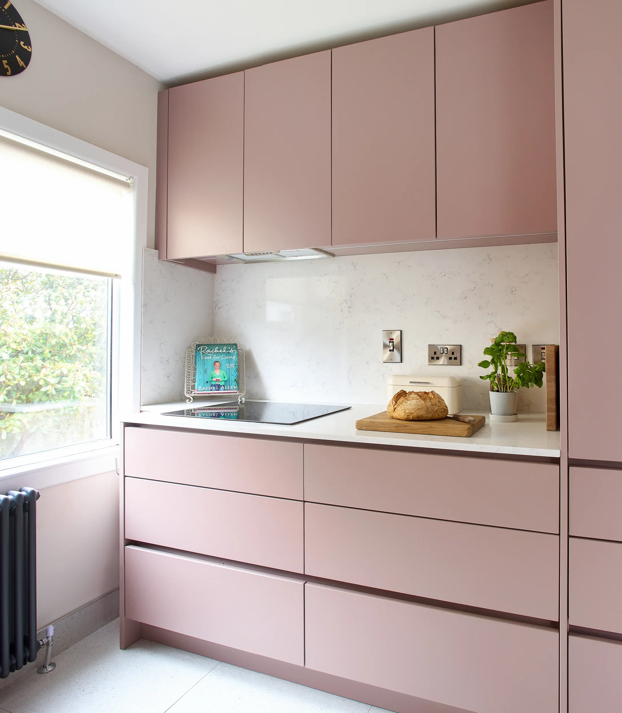 Modern kitchen with pink cabinets, white marble backsplash, window with roller blind, some plants, a stone, bread, and a book on the counter.